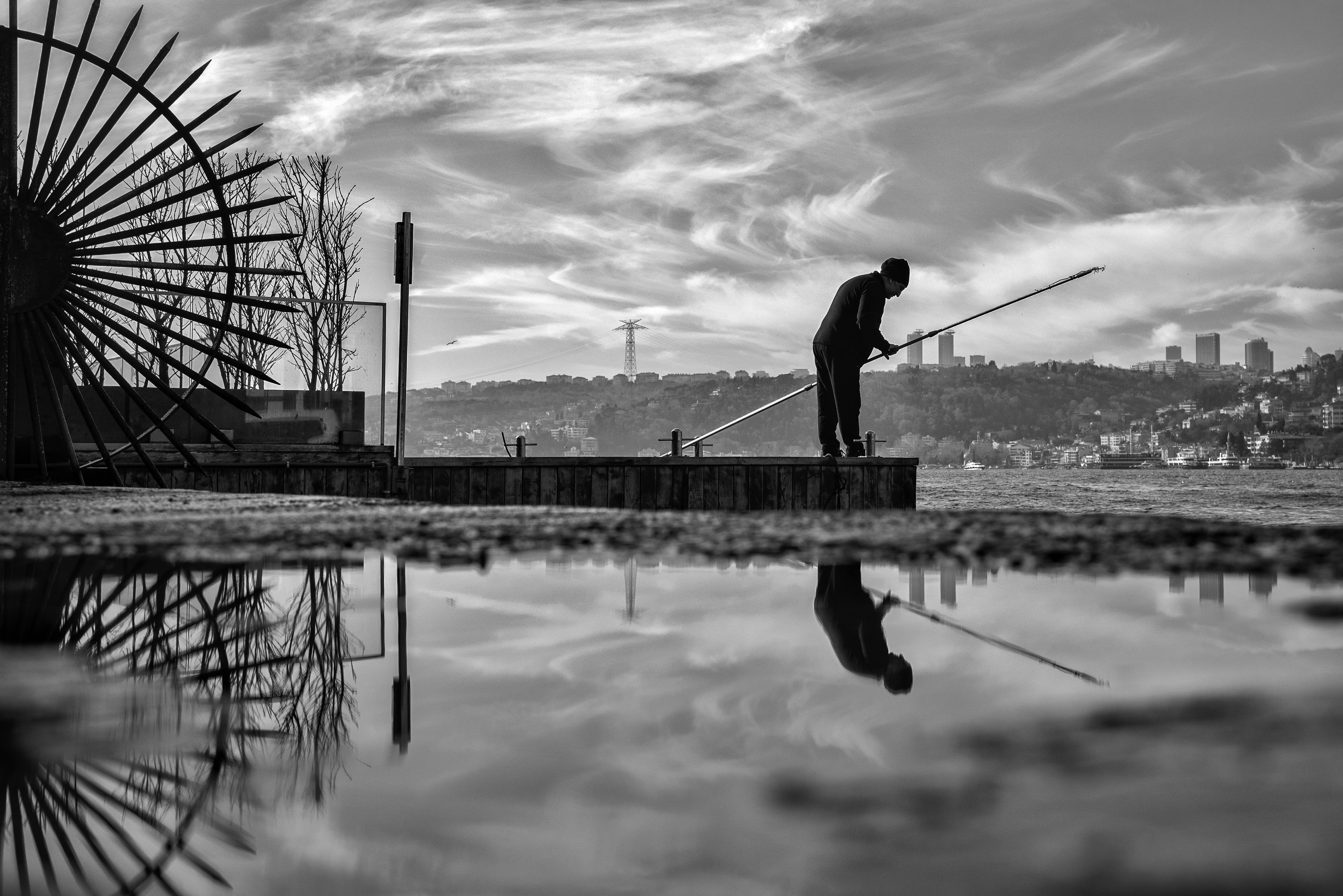 Black and white silhouette of a fisherman on a waterfront pier with a vivid sky reflection.