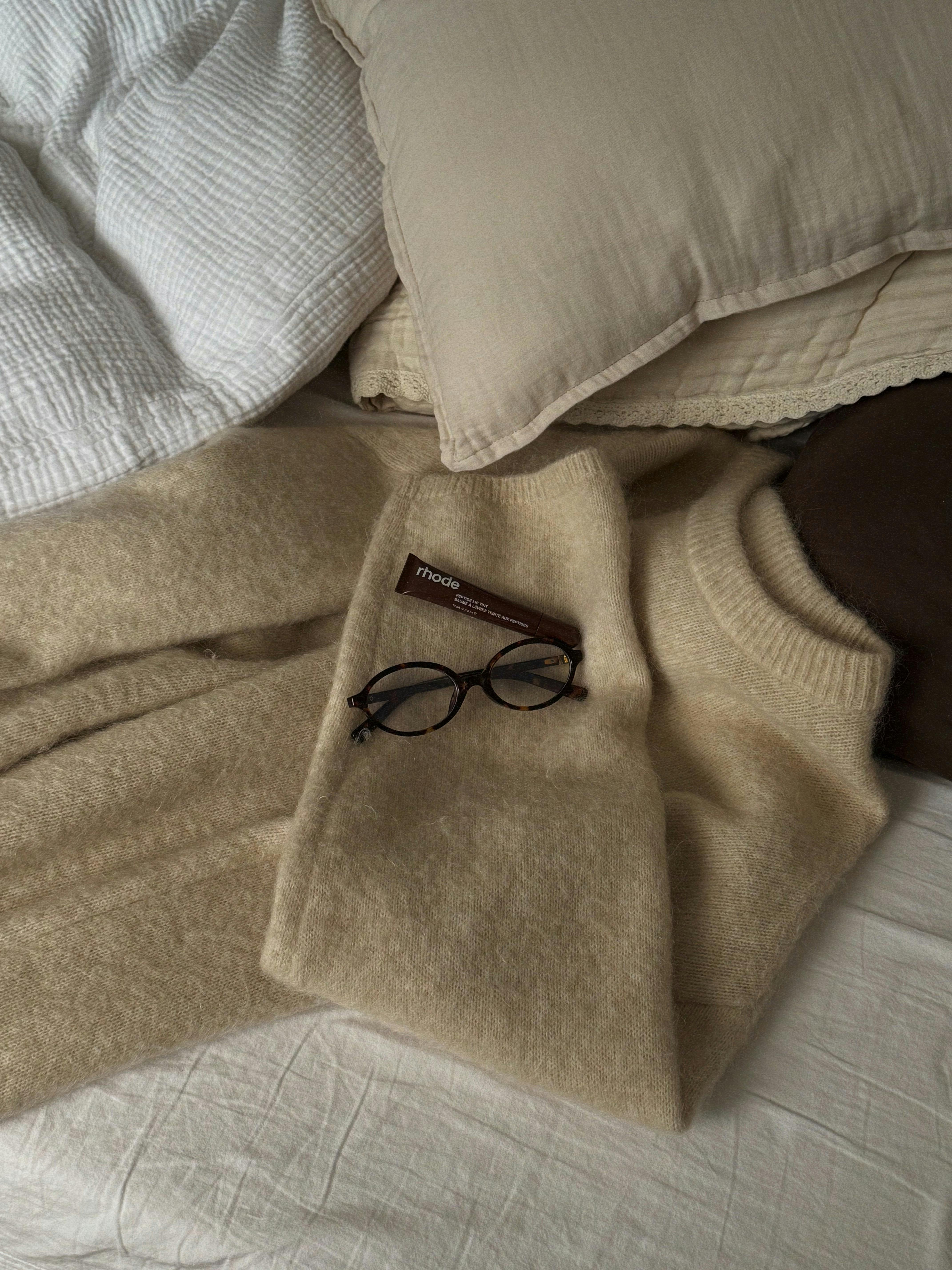 A soft beige sweater with glasses rests on a bed, exuding comfort and warmth.