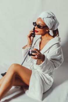 Chic woman in robe with sunglasses, talking on phone, enjoying leisure indoors.
