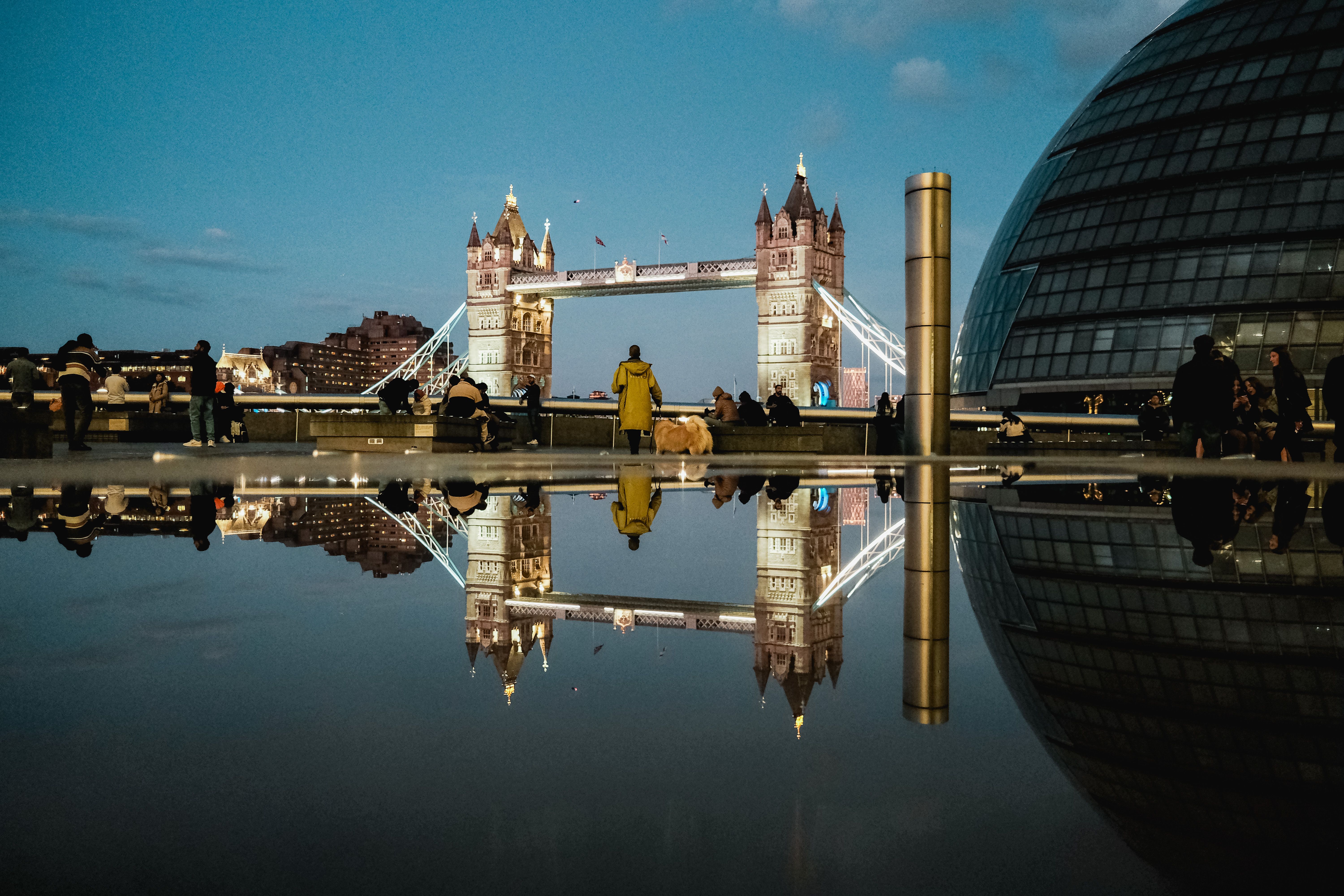 Night view of London's Tower Bridge reflected in water, surrounded by people and city lights.