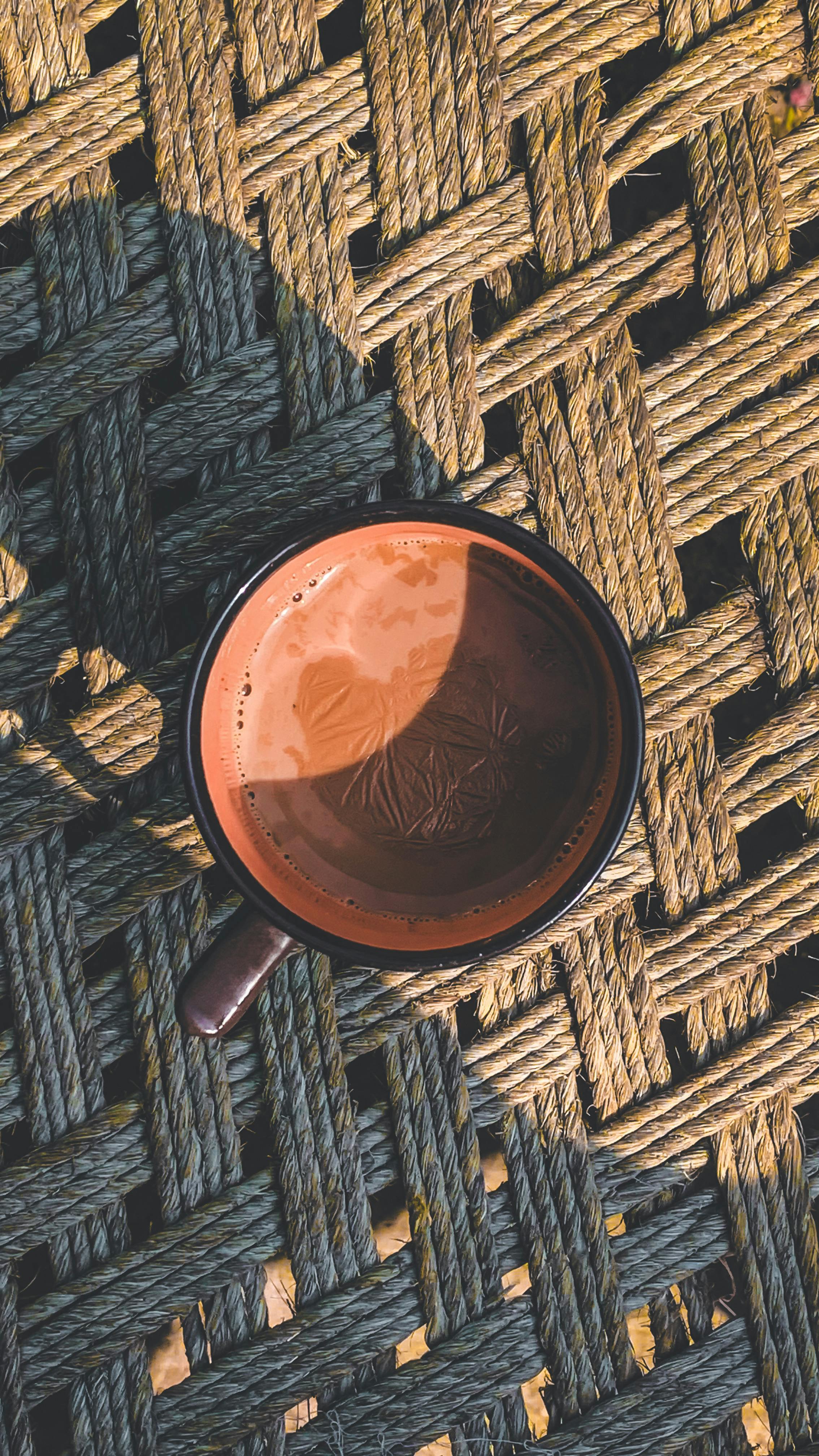 grátis Uma caneca de café marrom projetando uma sombra sobre um tapete de tecido sob a luz do sol. Foto profissional