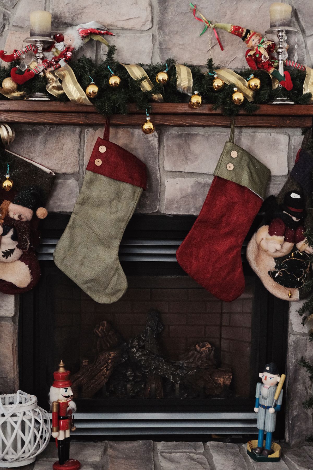 Sage green and red Christmas stockings hanging from a decorated fireplace mantel with garland and ornaments