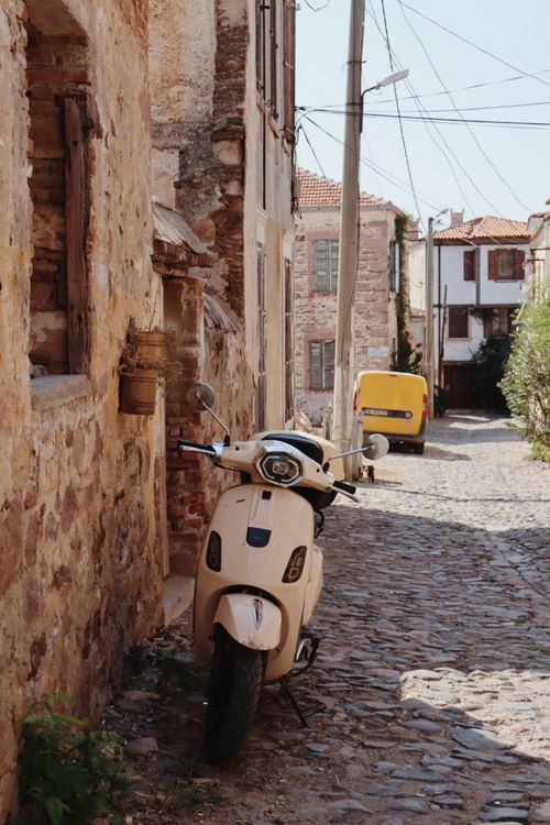 Free Vintage Vespa parked in a sunlit cobblestone alley surrounded by rustic buildings. Stock Photo
