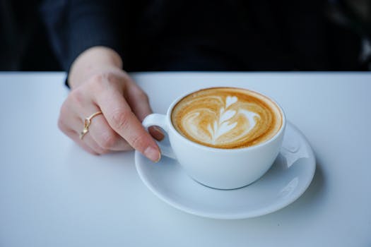Close-up of a latte with artistic foam, held by a human hand on a white table.