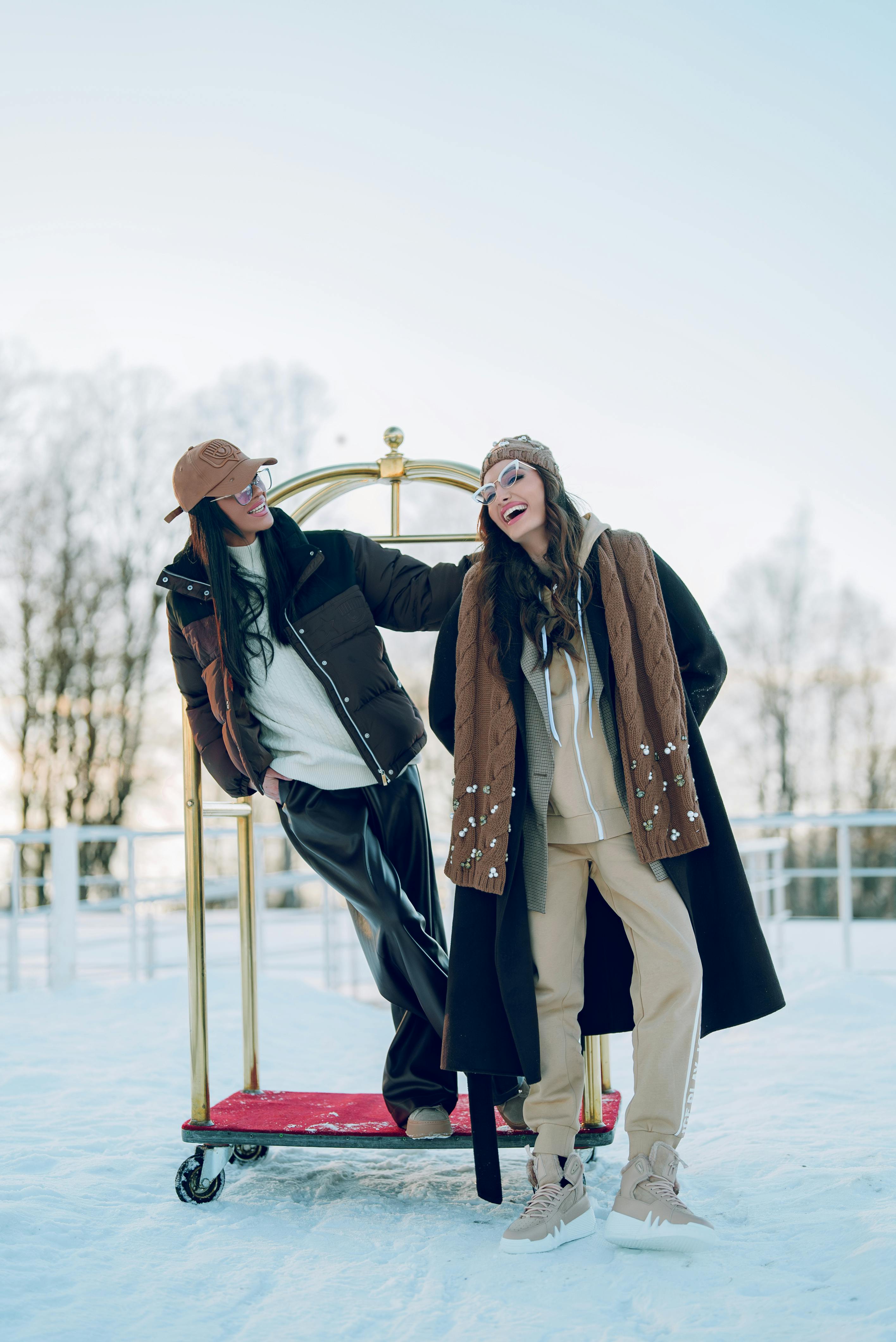 Two stylish women laughing in snowy winter scenery with a luggage cart, dressed warmly in winter fashion.
