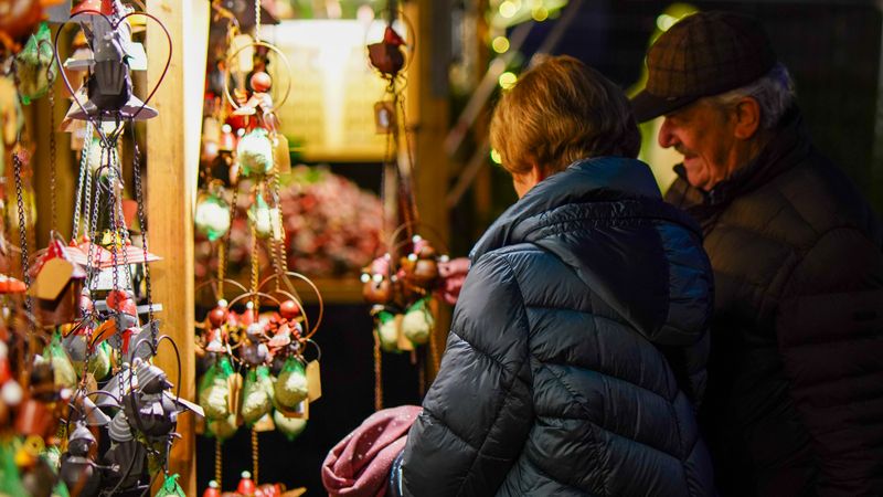 Älteres Paar in Winterjacken betrachtet abends an einem beleuchteten Marktstand hängenden Weihnachtsschmuck