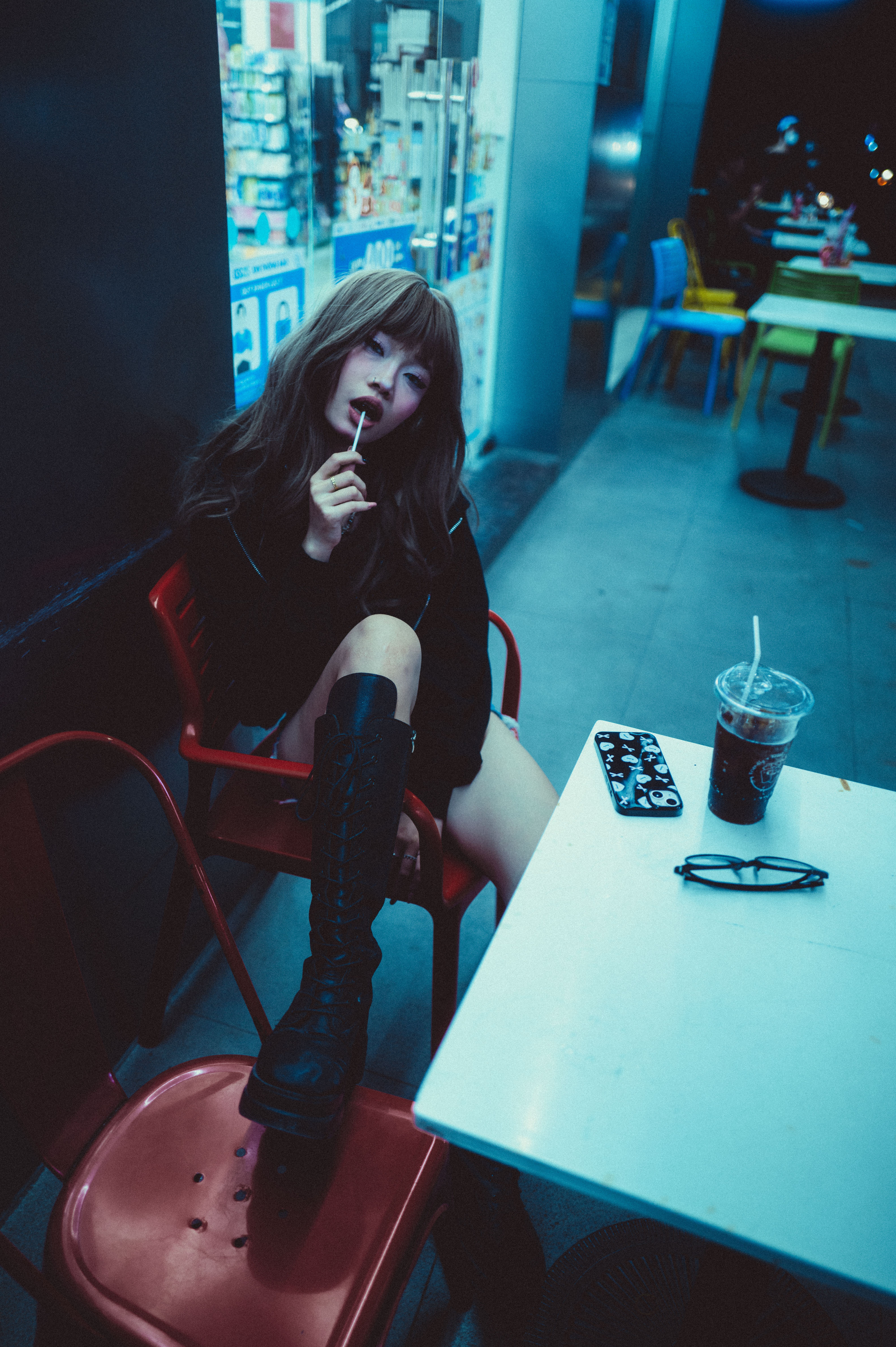 A stylish woman sitting outdoors at a cafe, enjoying a night scene with a drink and smartphone.