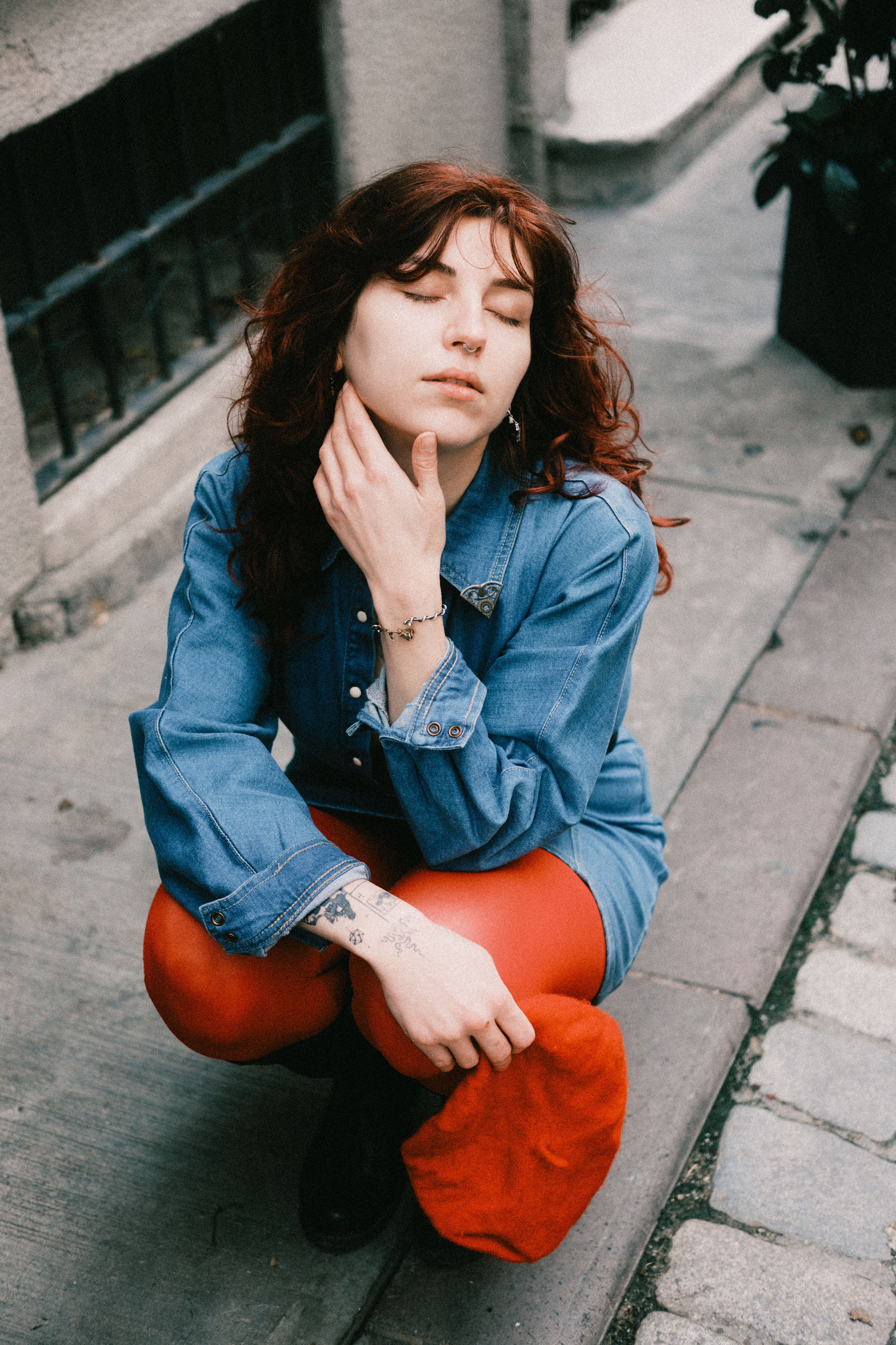 A stylish young woman squats outdoors, wearing red tights and a denim jacket.