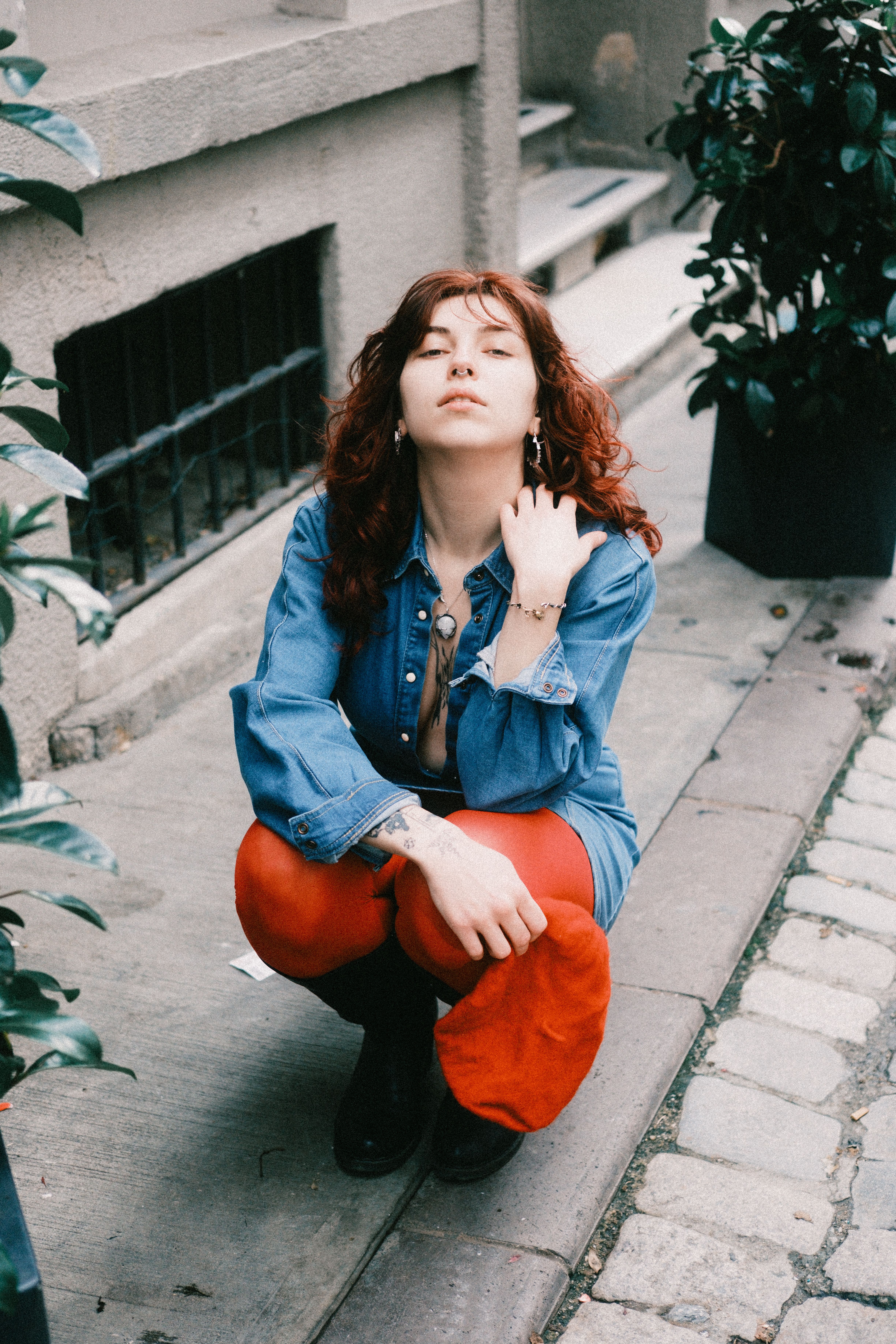 Red-haired woman in denim jacket and red pants poses in a modern urban setting.