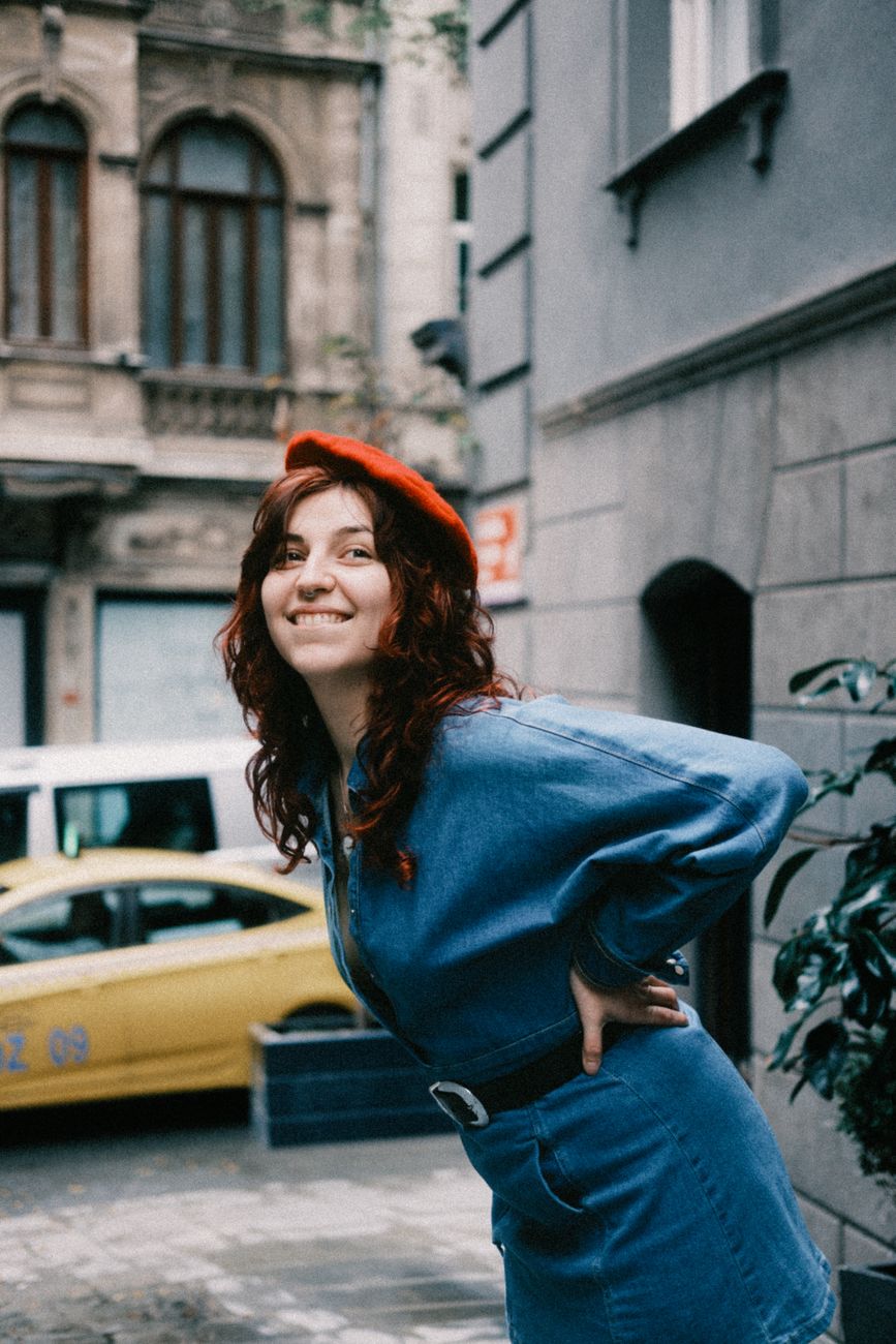Smiling woman in denim and red beret poses on a city street with vintage architecture.