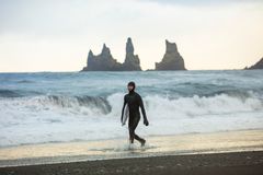 Un surfista camina sobre las arenas negras de la playa Reynisfjara, Islandia, sosteniendo una tabla de surf.