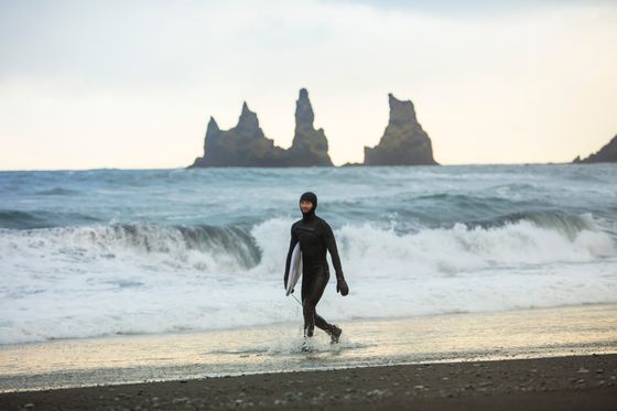 Bir sörf tahtası tutan bir sörfçü, İzlanda'daki Reynisfjara Plajı'nın siyah kumlarında yürüyor.