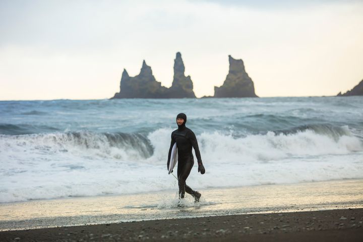A surfer walks on the black sands of Reynisfjara Beach, Iceland, holding a surfboard.