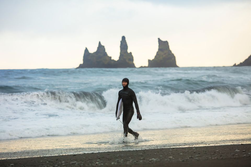 A surfer walks on the black sands of Reynisfjara Beach, Iceland, holding a surfboard.