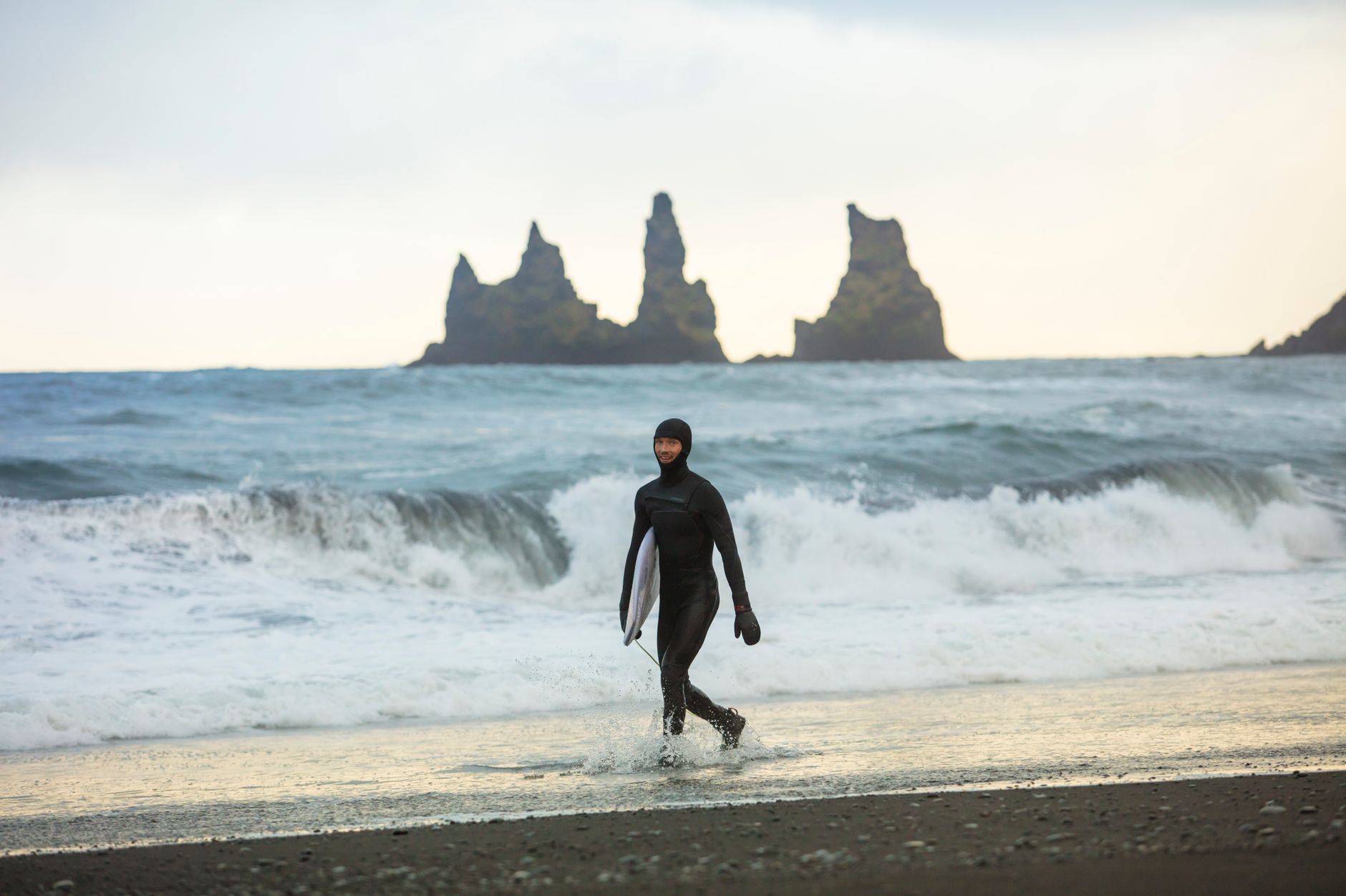 Un surfista camina sobre las arenas negras de la playa Reynisfjara, Islandia, sosteniendo una tabla de surf.