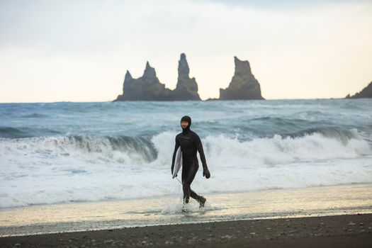 A surfer walks on the black sands of Reynisfjara Beach, Iceland, holding a surfboard.