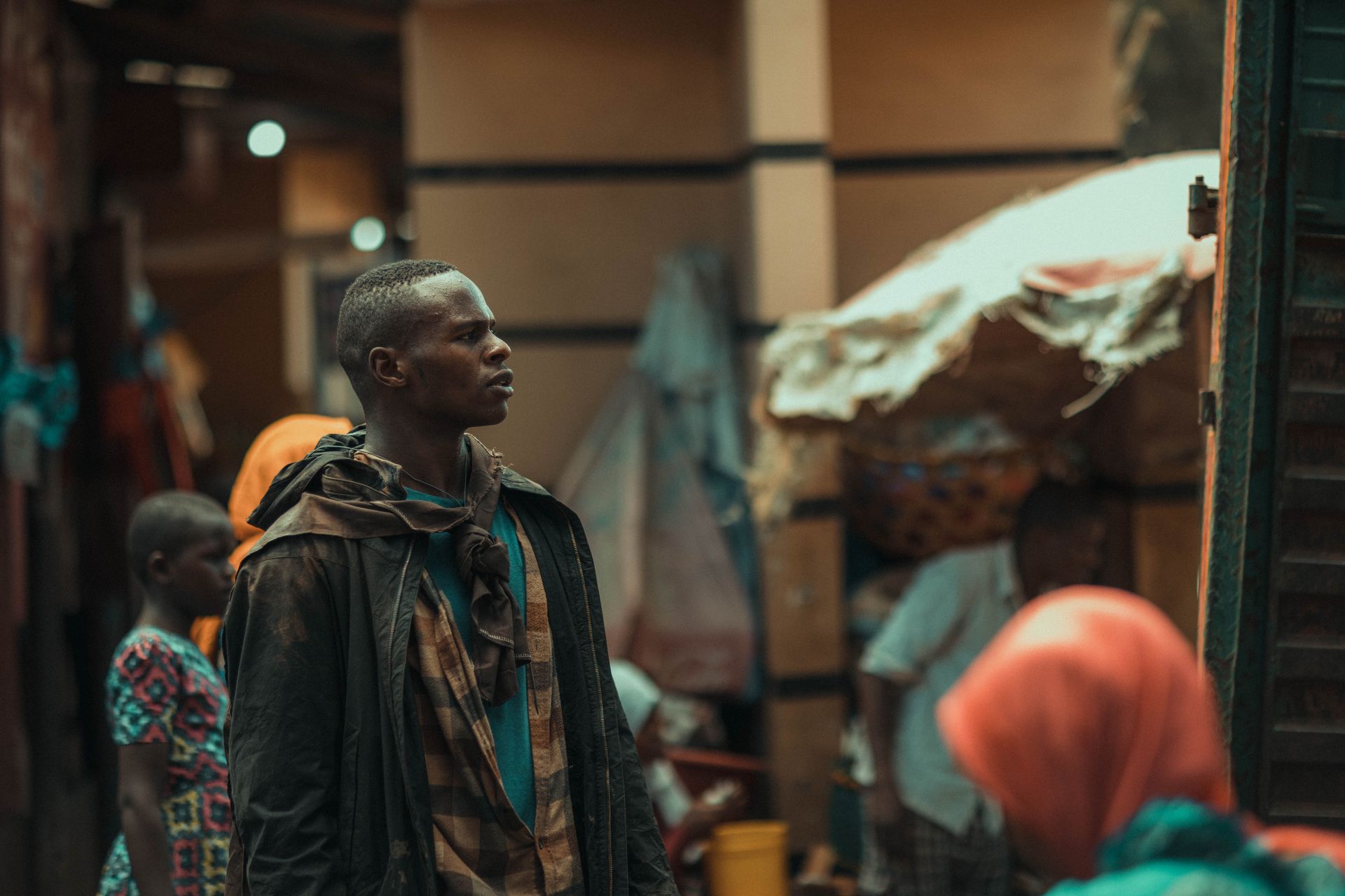 A young man stands in an urban market surrounded by vibrant colors and people.