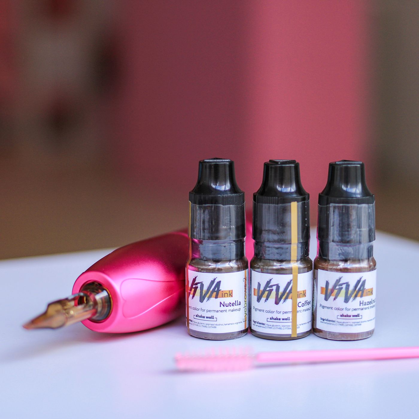 Three permanent makeup pigment bottles displayed beside a pink cosmetic tattoo machine and tools.
