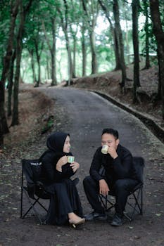 Couple enjoying hot drinks in chairs amidst a serene forest path.
