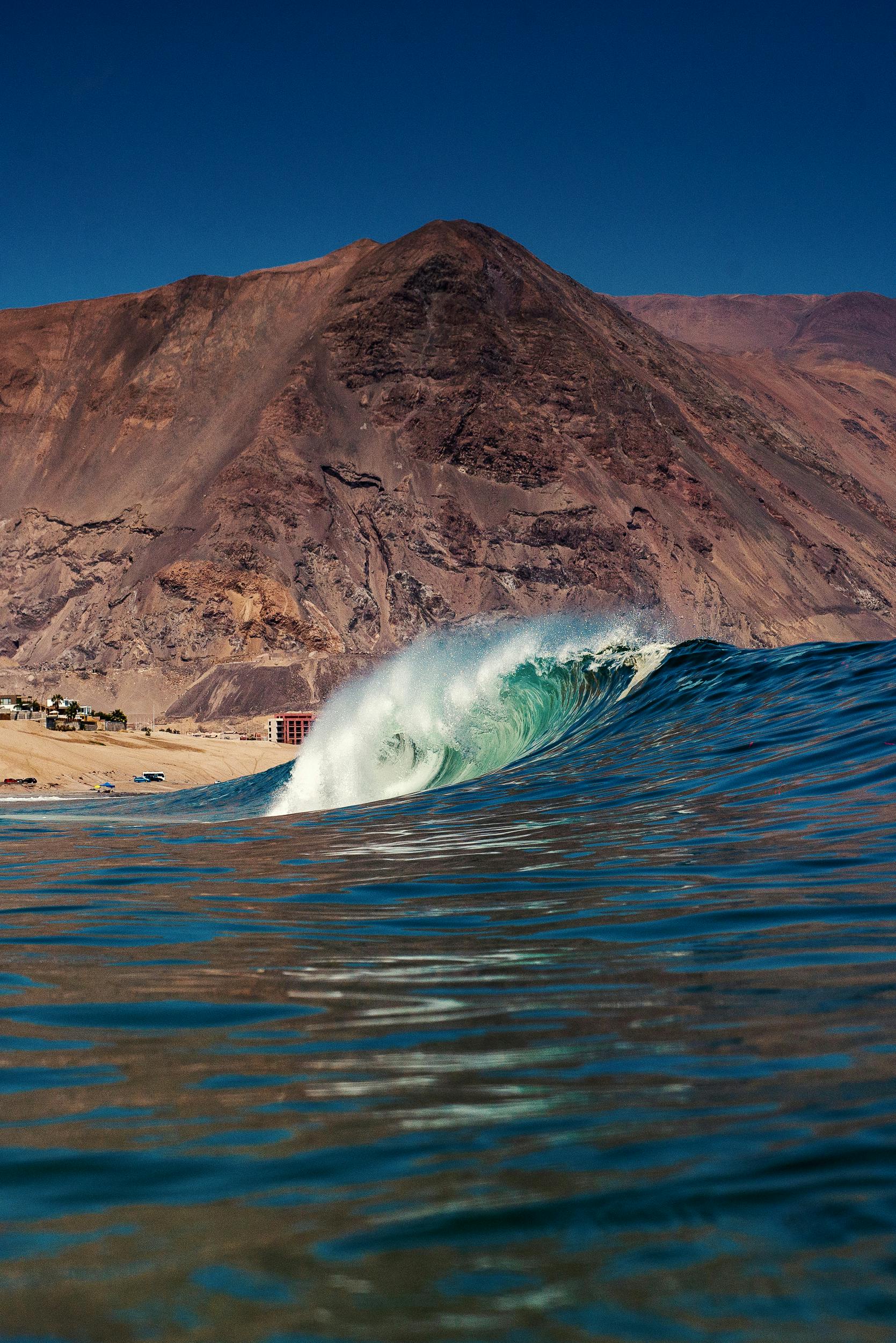A stunning wave crashes on the beach with the backdrop of Iquique's majestic mountains and clear skies.