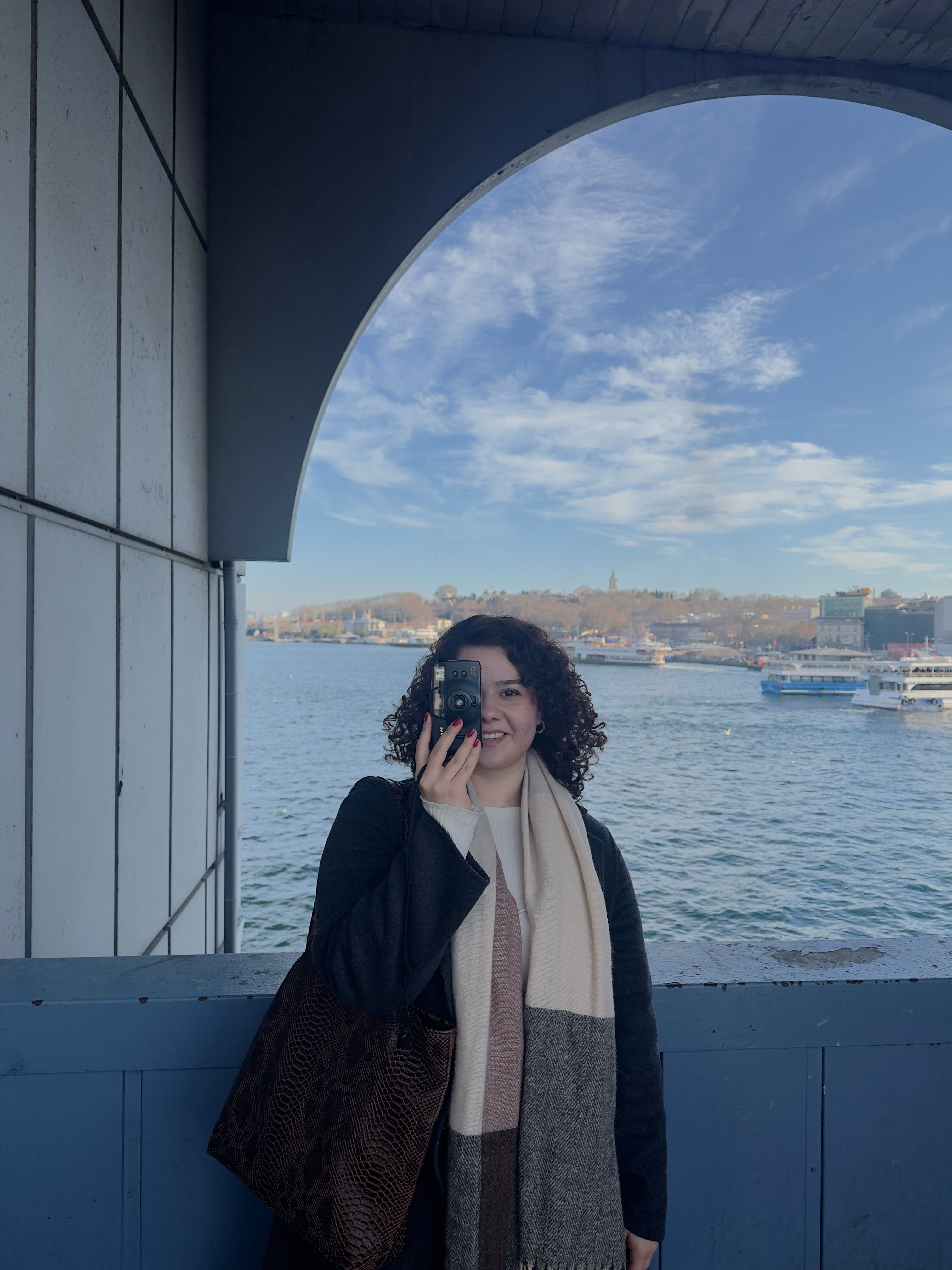 Smiling woman with camera at scenic waterfront beneath arch, capturing vibrant scenery.