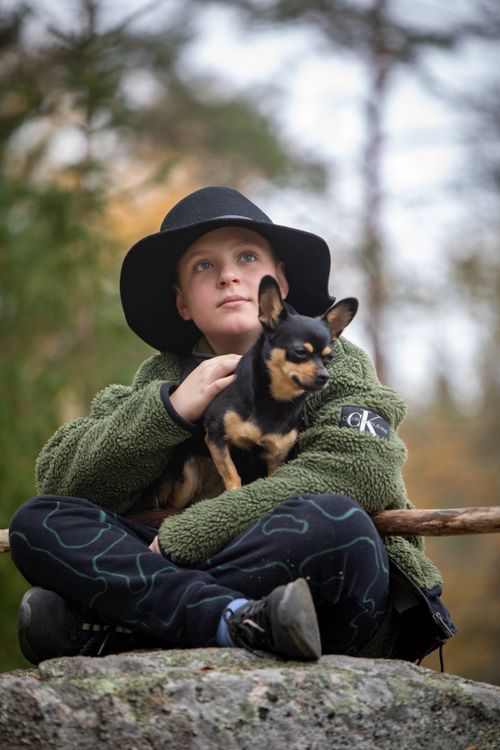 Free Young boy sitting in nature holding a Chihuahua, wearing a green jacket and black hat. Stock Photo