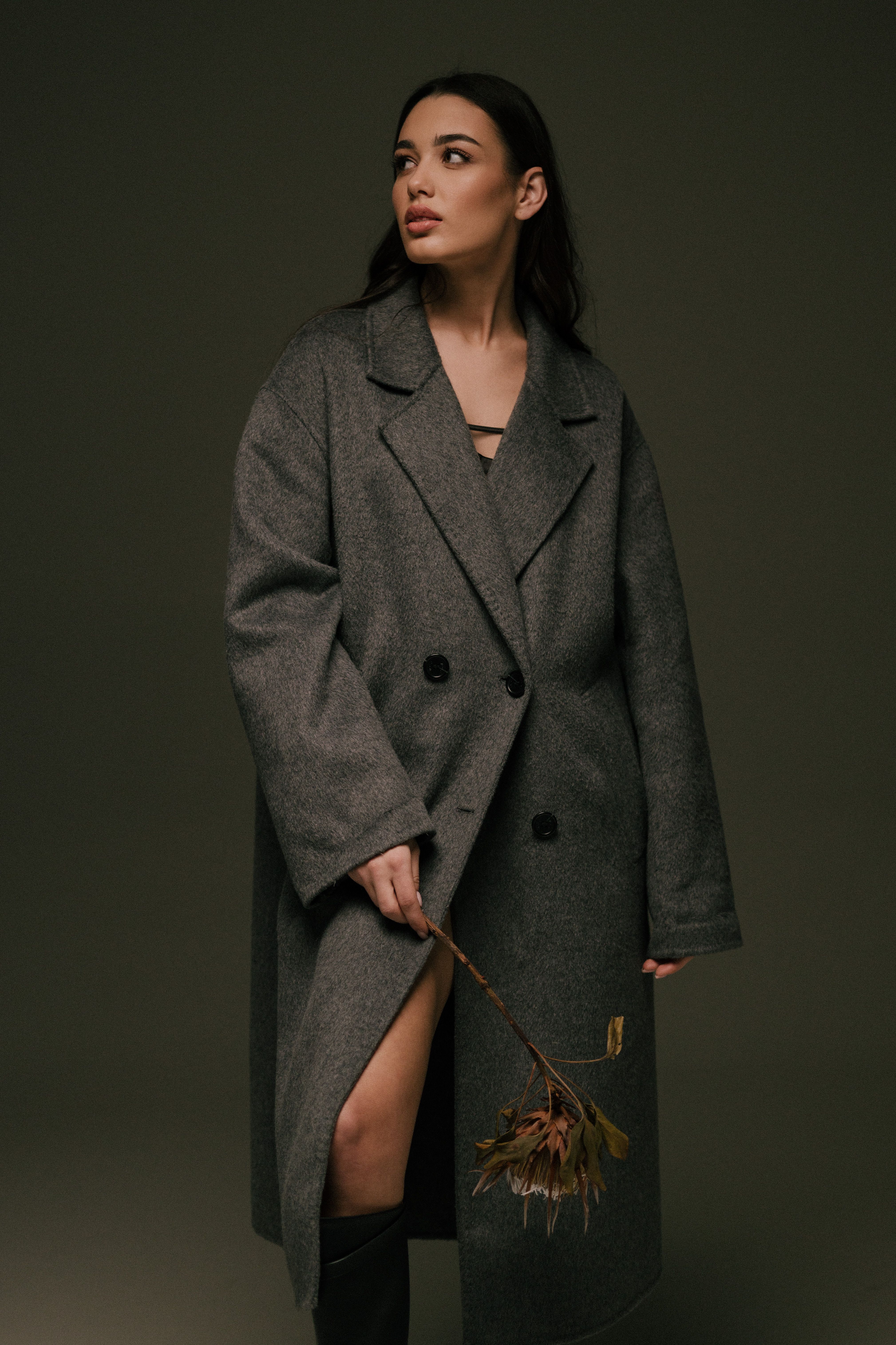Elegant woman in a grey coat holding a flower, captured in a moody studio setting.