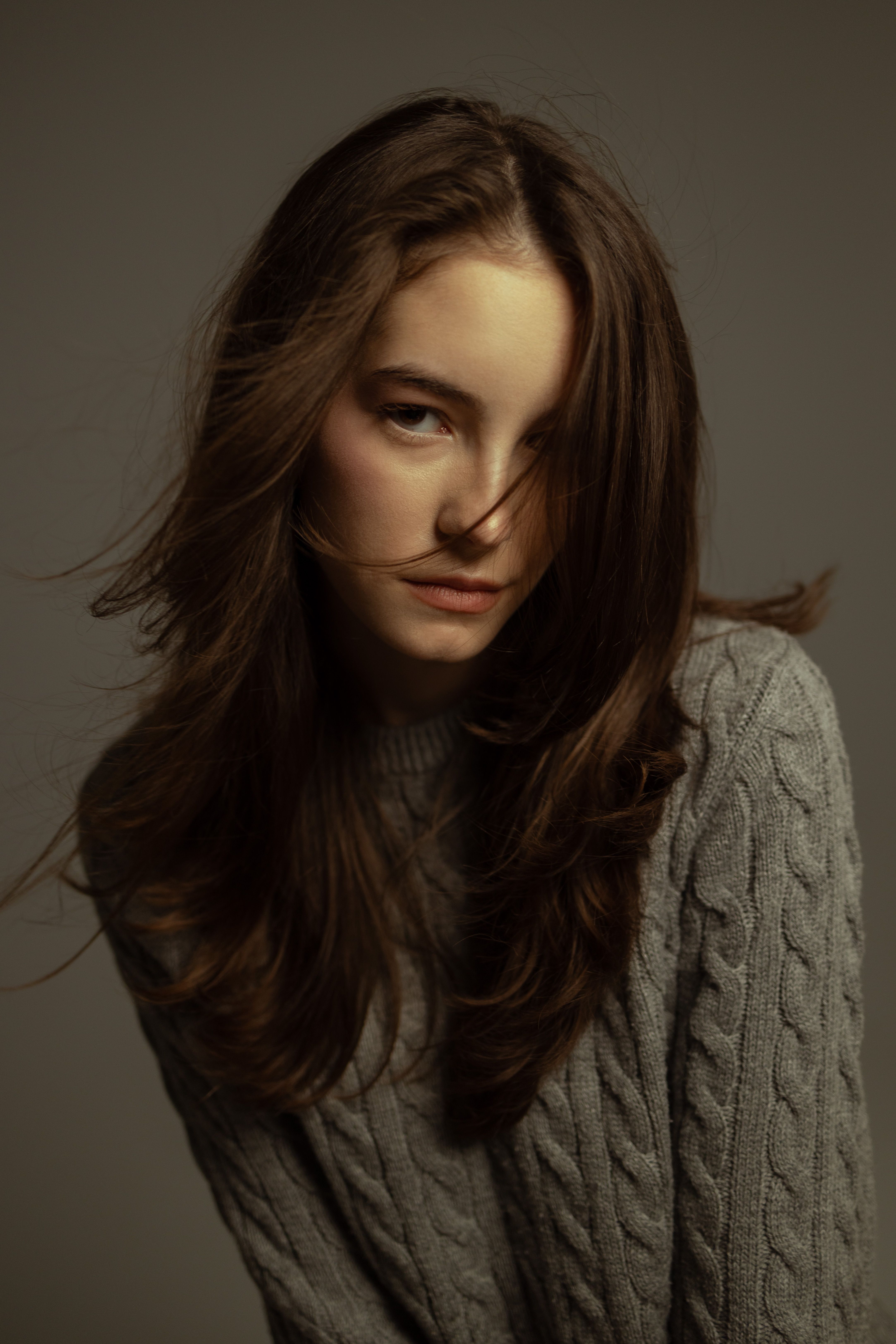 Stylish portrait of a woman with long hair wearing a grey sweater against a soft backdrop.