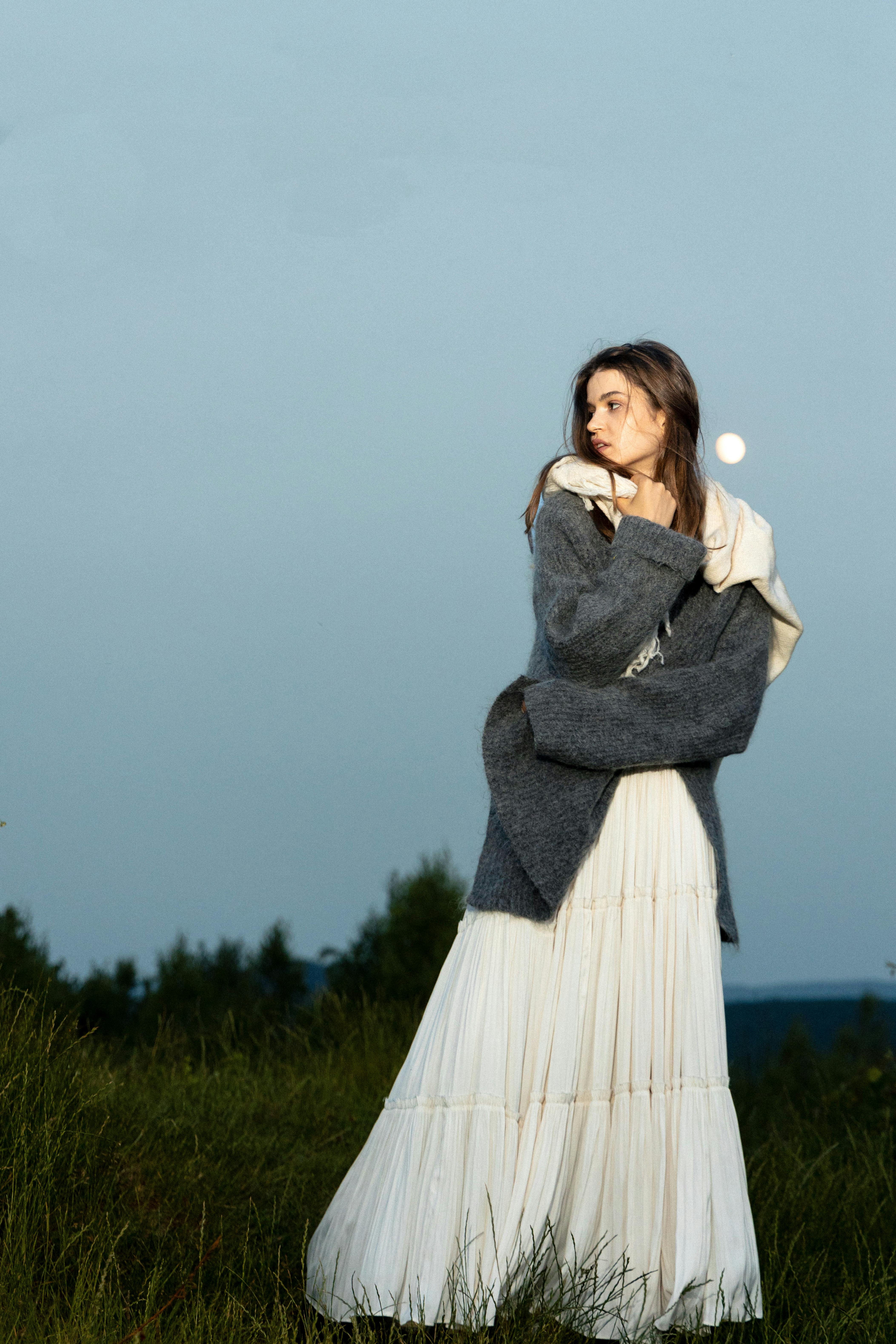 Elegant woman in long dress and coat outdoors under a moonlit sky, exuding a serene and contemplative mood.