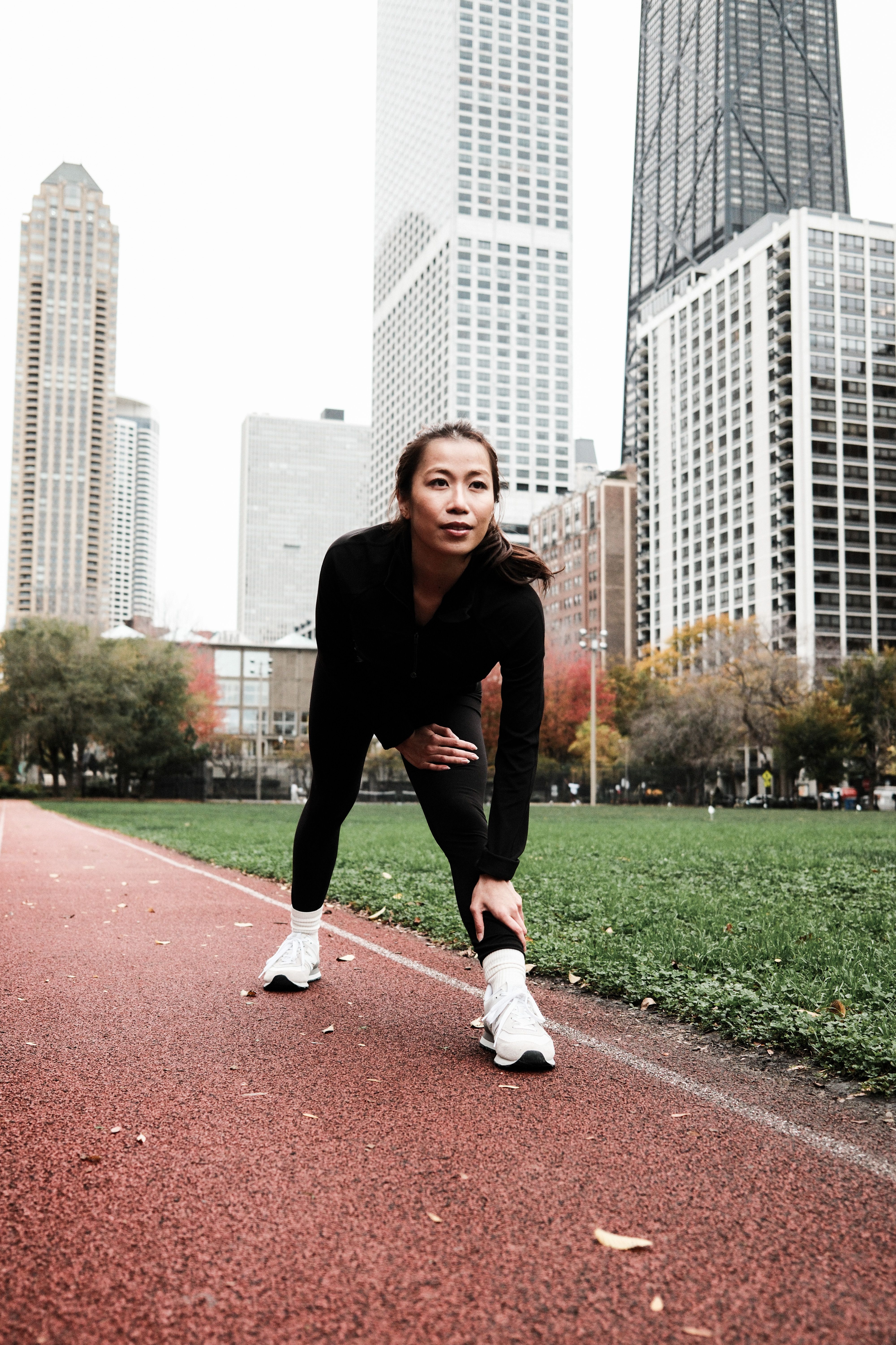 Asian woman in athletic wear stretching on a track in Chicago with a city skyline backdrop.