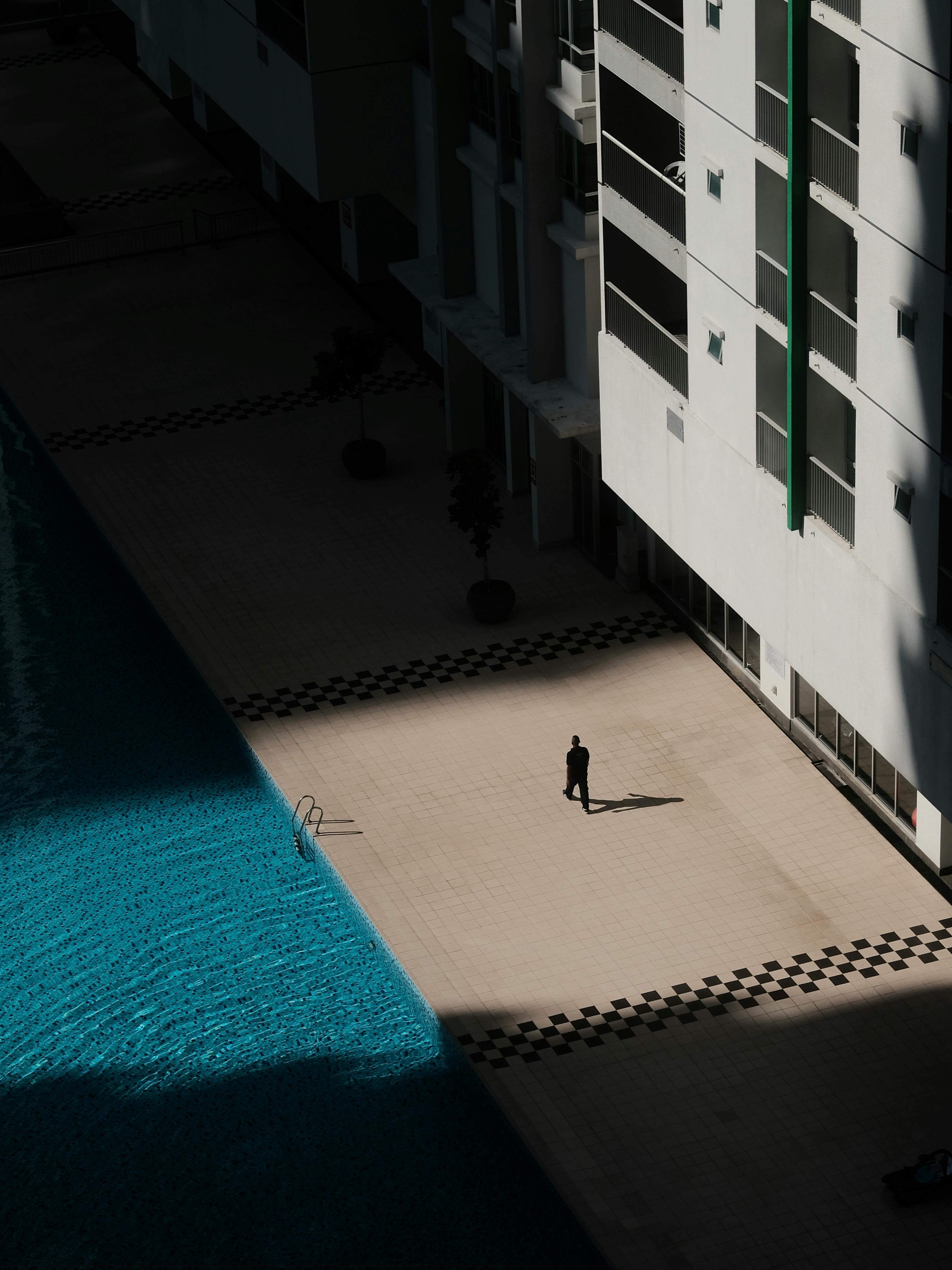 A lone figure casts a shadow by a bright blue poolside in a modern architectural setting.