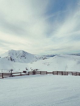A tranquil winter scene showcasing snow-covered mountains and a wooden fence under a vast blue sky.