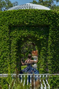 A woman in a blue dress stands in a lush garden gazebo surrounded by greenery.