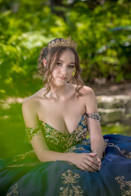 Free A young woman wearing a gold crown and ornate gown sits gracefully outdoors amid lush greenery. Stock Photo