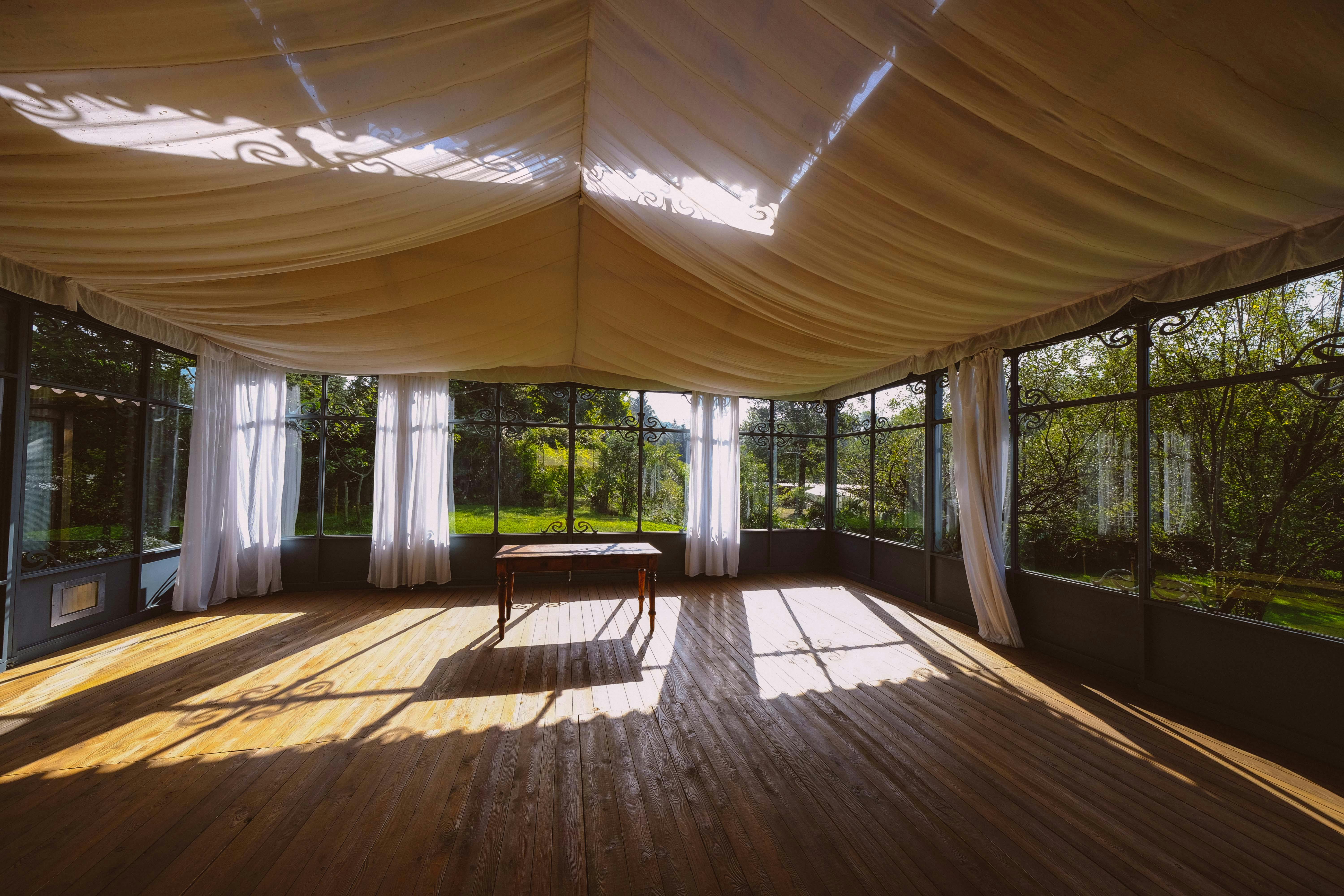 A bright conservatory room with wooden floors and a single table, offering a view of lush greenery outside.