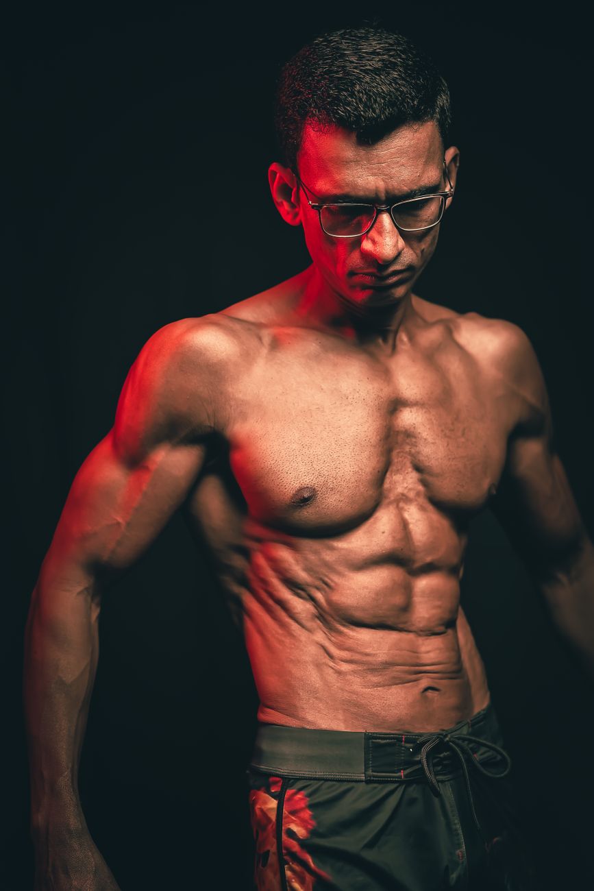 Fit man showcasing muscular physique with glasses, dramatic lighting.