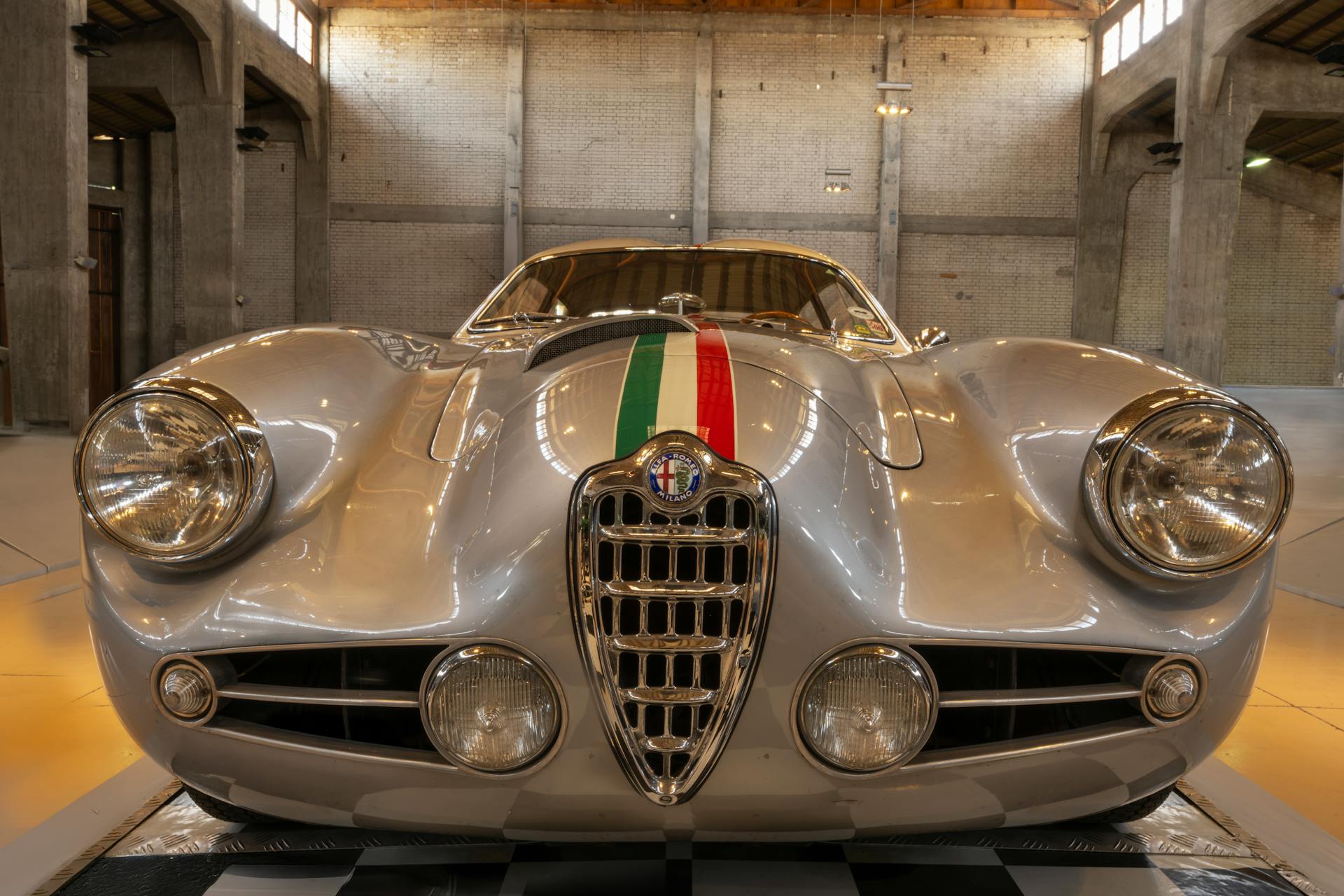 Front view of a vintage silver Alfa Romeo car inside an industrial garage setting.