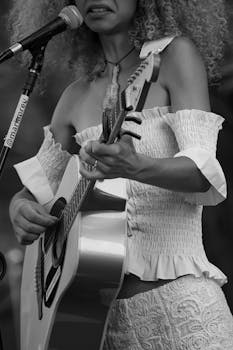 Monochrome image of a woman playing acoustic guitar on stage, showcasing her curly hair and off-shoulder attire.