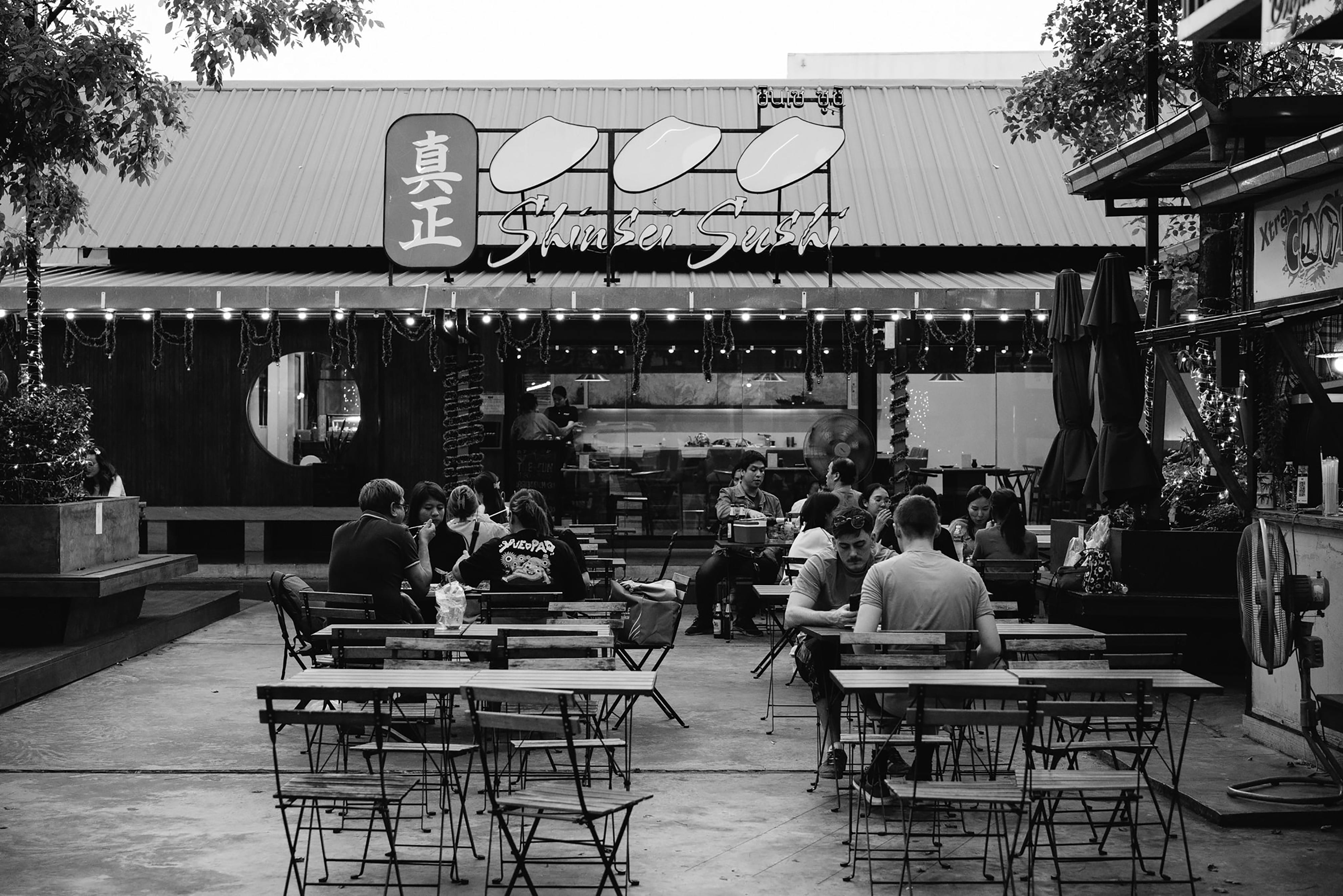A lively outdoor dining scene at a sushi restaurant captured in black and white, showcasing customers enjoying their meals.