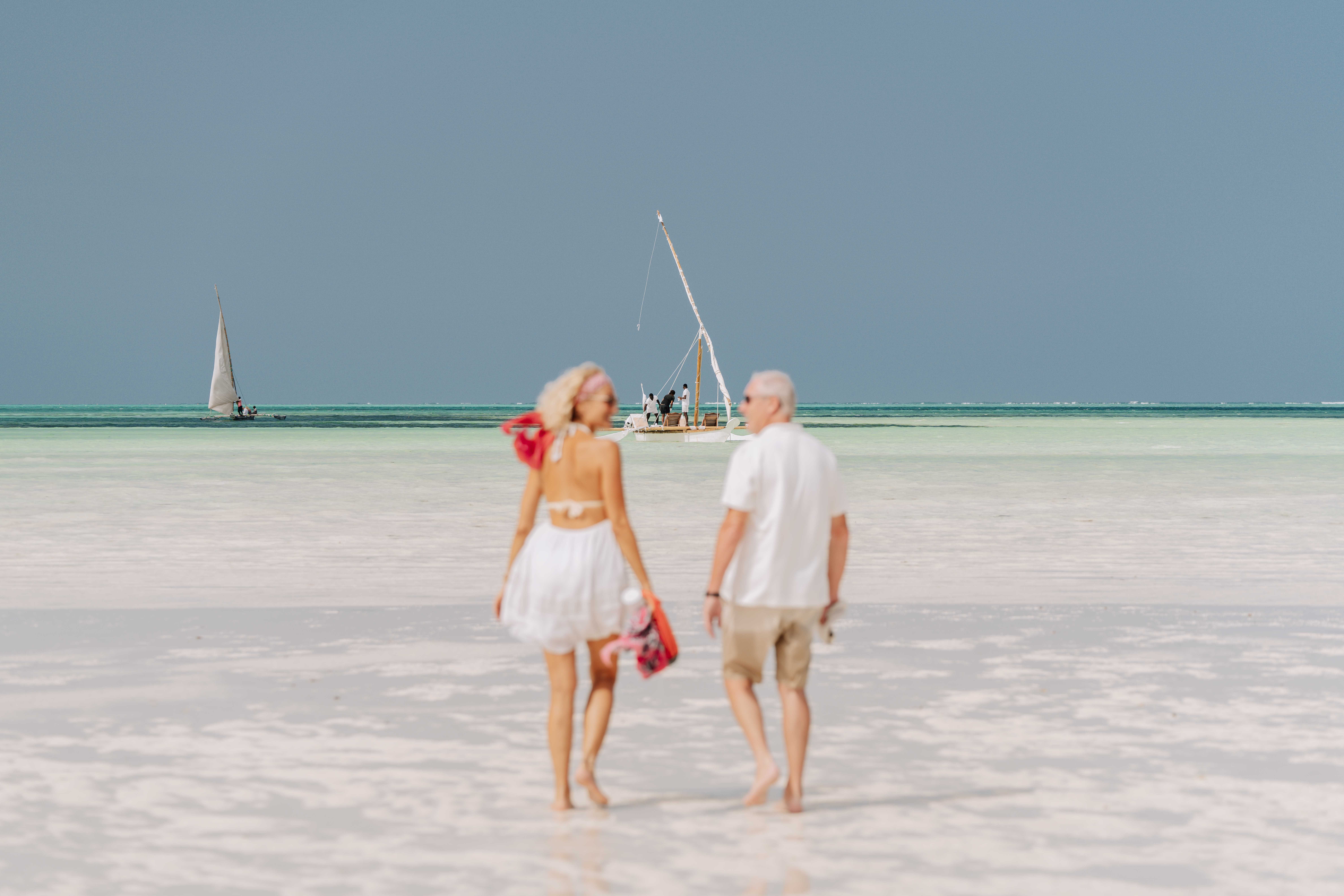 Elderly couple enjoying a serene walk on a sunny Zanzibar beach with visible dhows. Elderly couple enjoying a serene walk on a sunny Zanzibar beach with visible dhows.