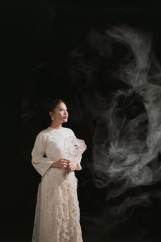 A poised woman in a long vintage dress gazing against a dark smoky backdrop.