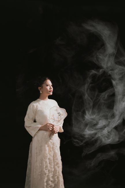 A poised woman in a long vintage dress gazing against a dark smoky backdrop.