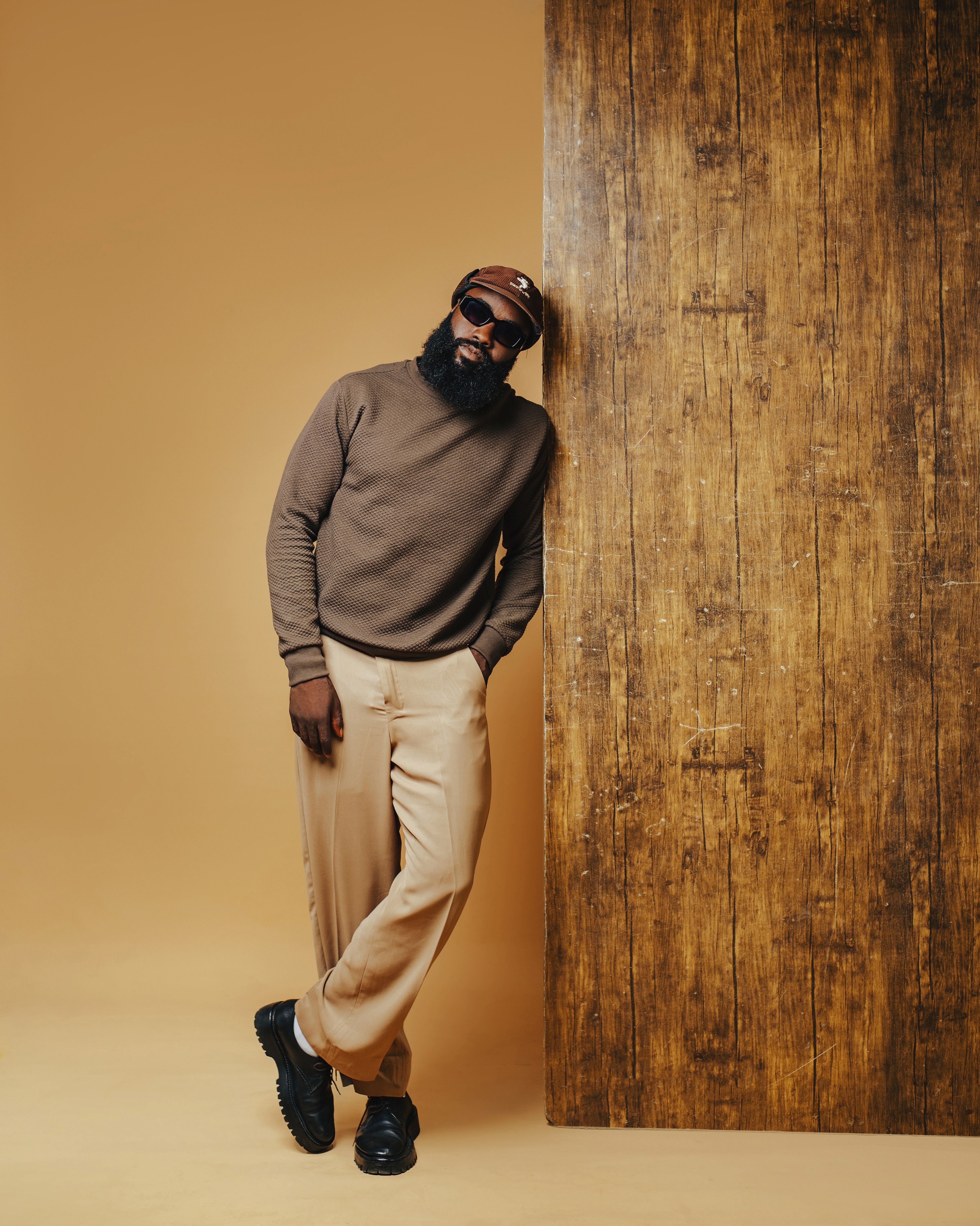 Portrait of a man in a brown sweater and beige pants leaning on a wooden wall.