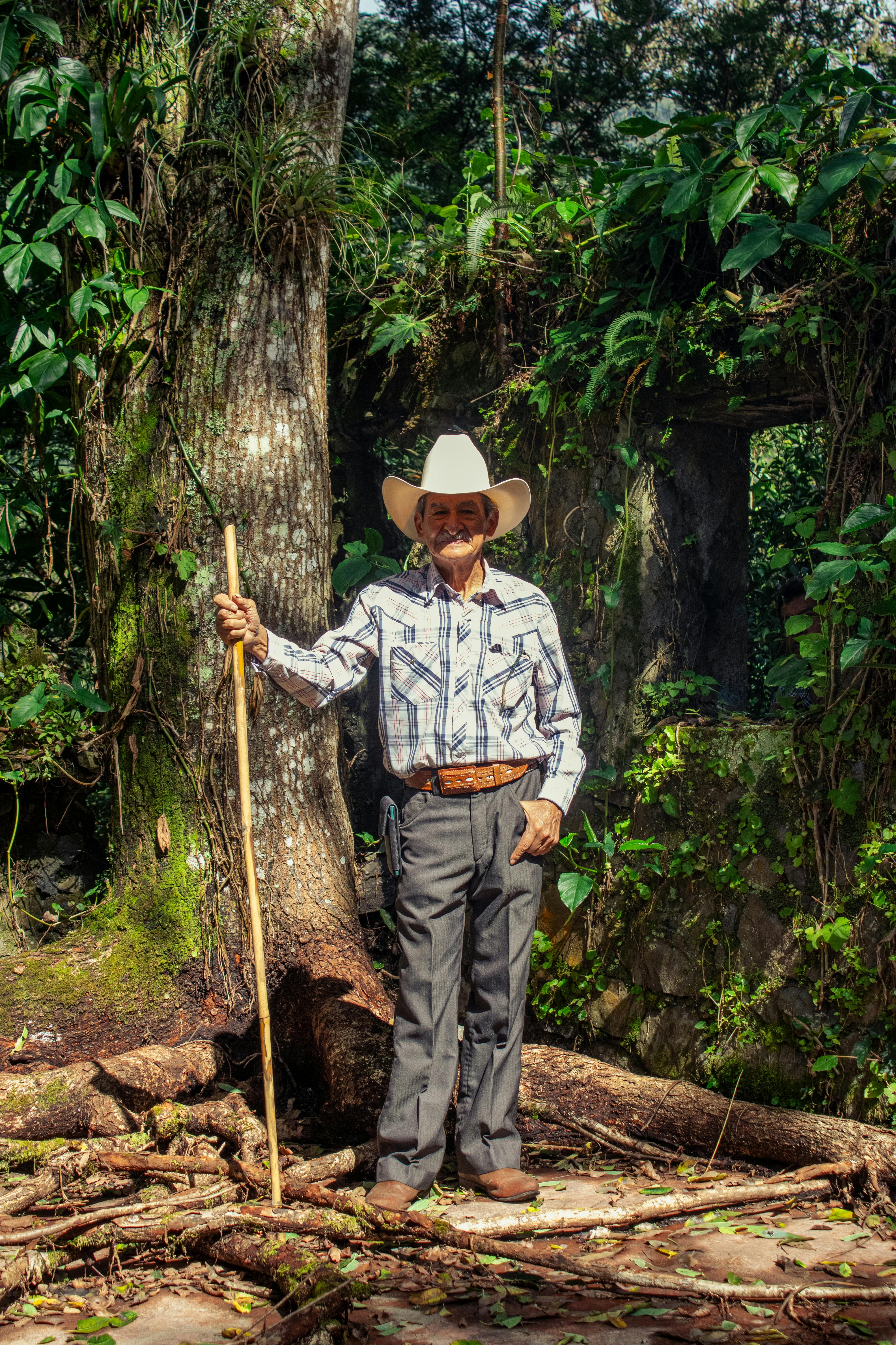 Elderly man in a white cowboy hat holding a stick amidst a lush green forest backdrop.