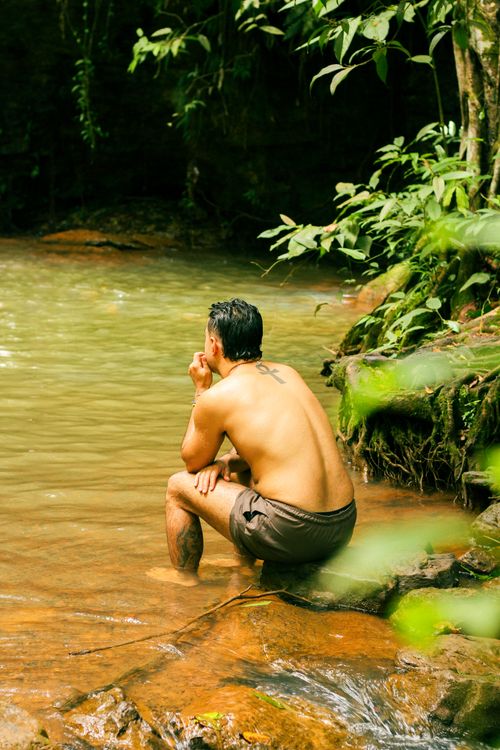 Free A man sits thoughtfully by a serene forest stream, surrounded by nature. Stock Photo