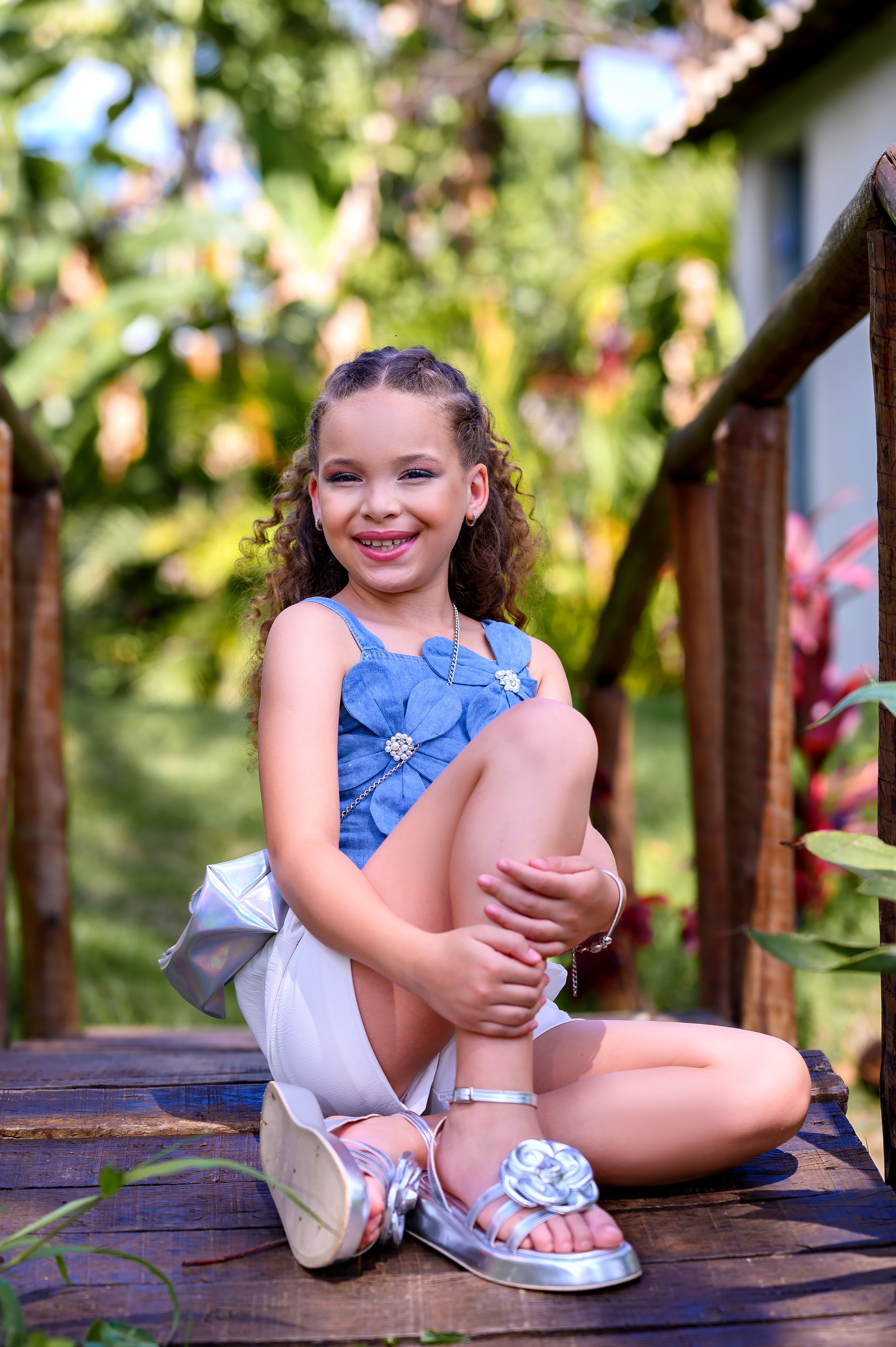 Smiling girl in summer attire sitting on a rustic wooden bridge surrounded by greenery.