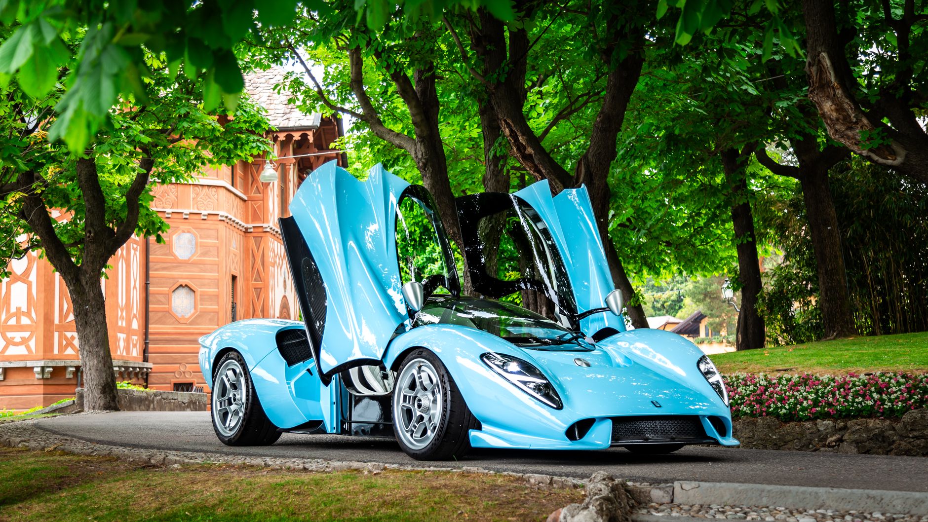 Striking blue sports car with butterfly doors open amidst lush greenery in a park.