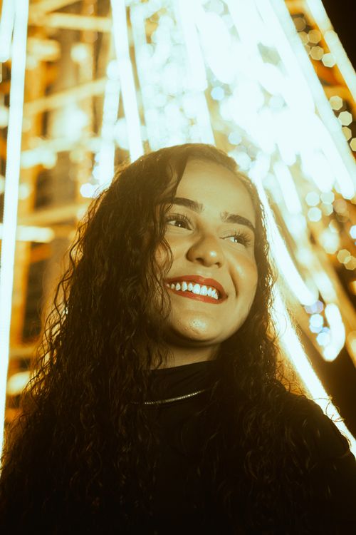 Free Happy young woman with curly hair smiling under bright festival lights at night. Stock Photo
