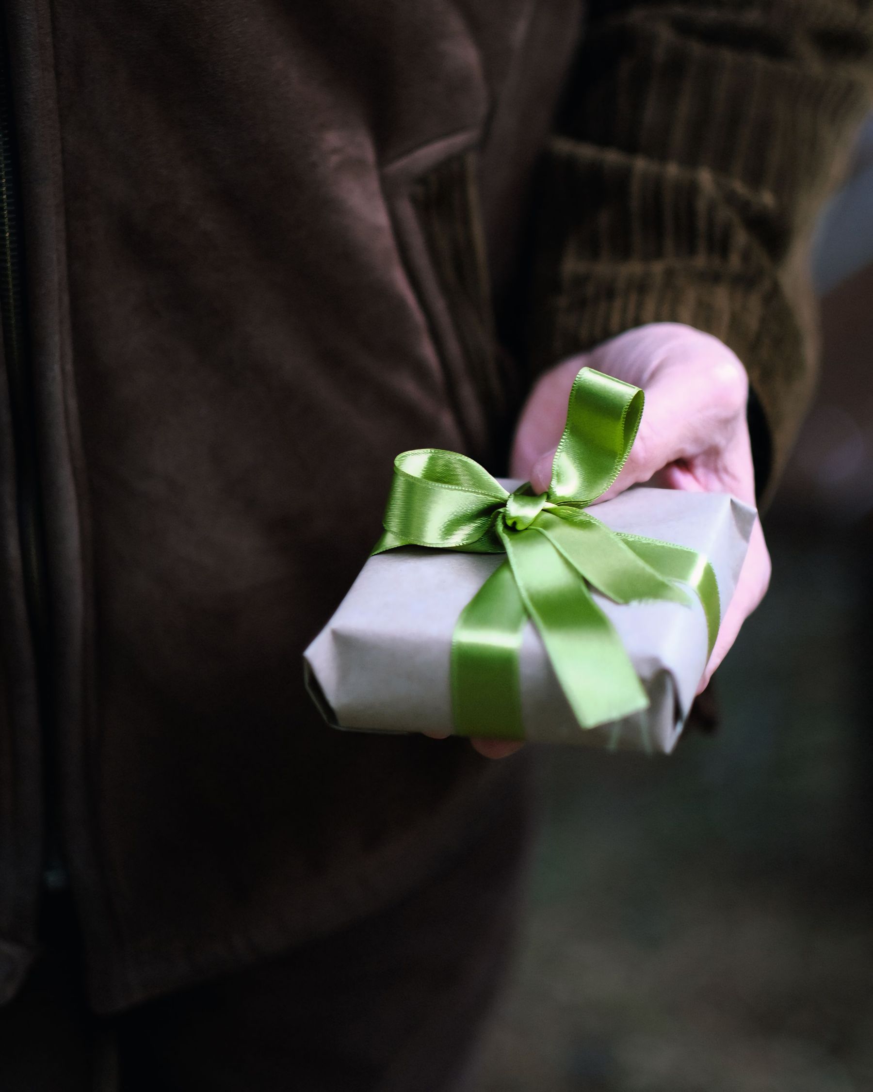 Person in a dark coat holding out a small wrapped gift with a green bow