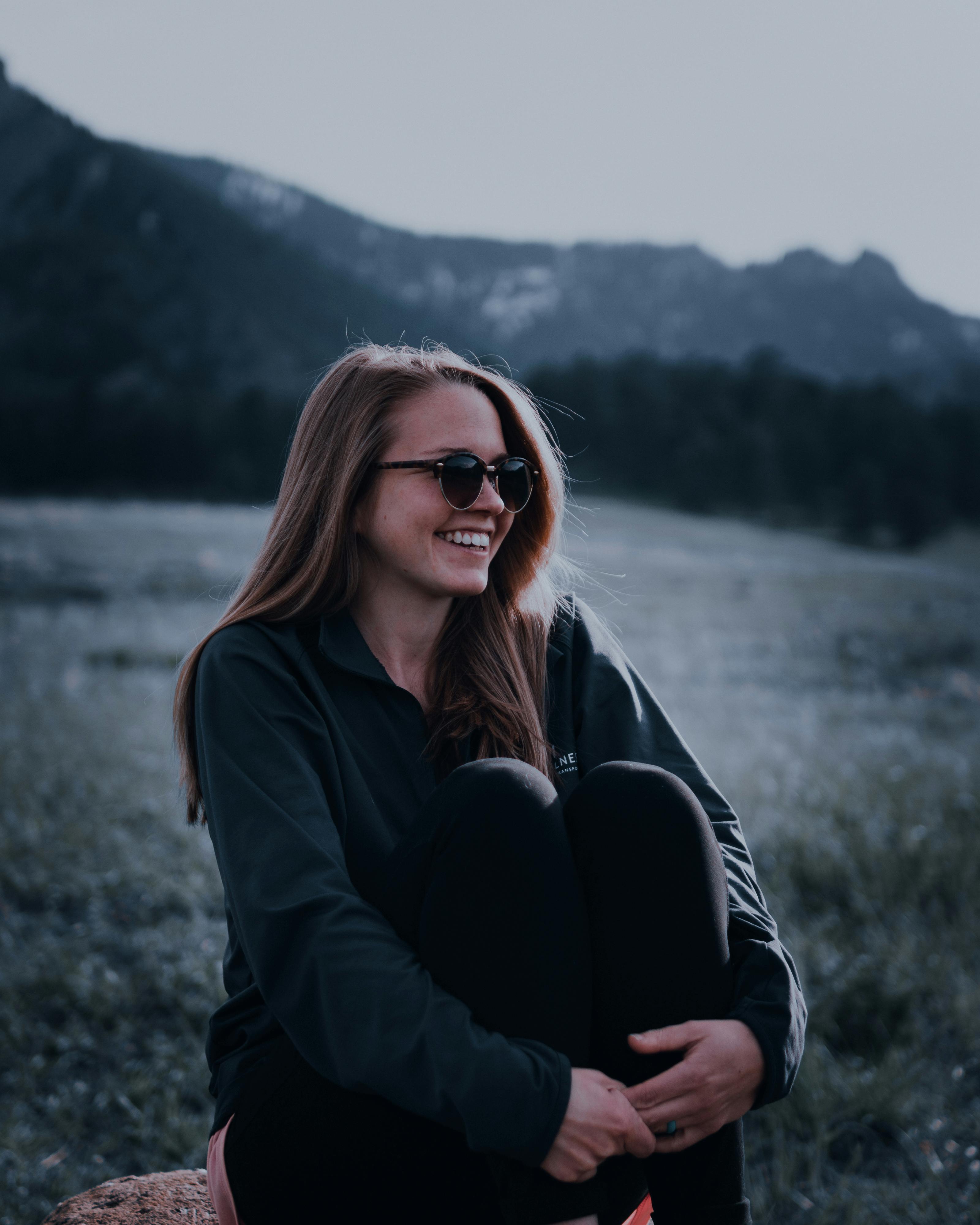 Happy woman with sunglasses sitting outdoors in a field with a mountain backdrop, embracing nature's serenity.